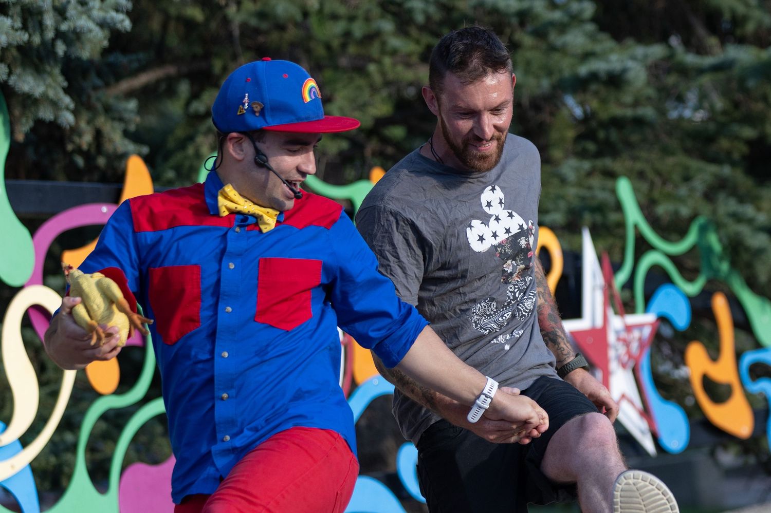 Colorfully dressed clown entertains man in gray shirt at outdoor festival with graffiti backdrop.
