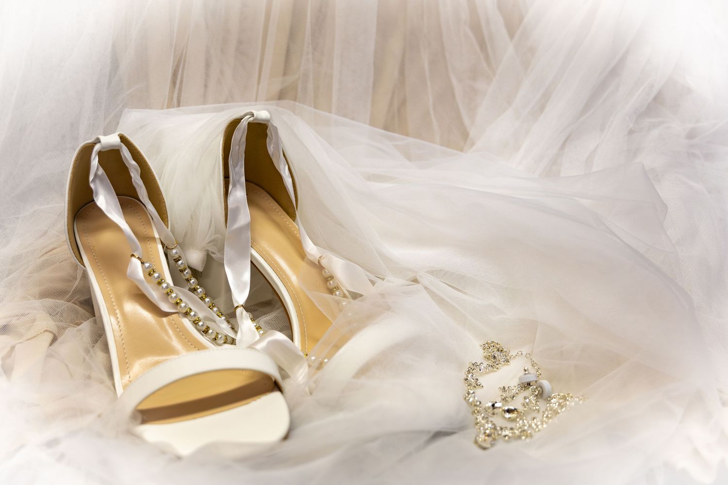 Elegant white bridal heels with crystal embellishments rest on a flowing wedding veil beside a sparkling brooch.