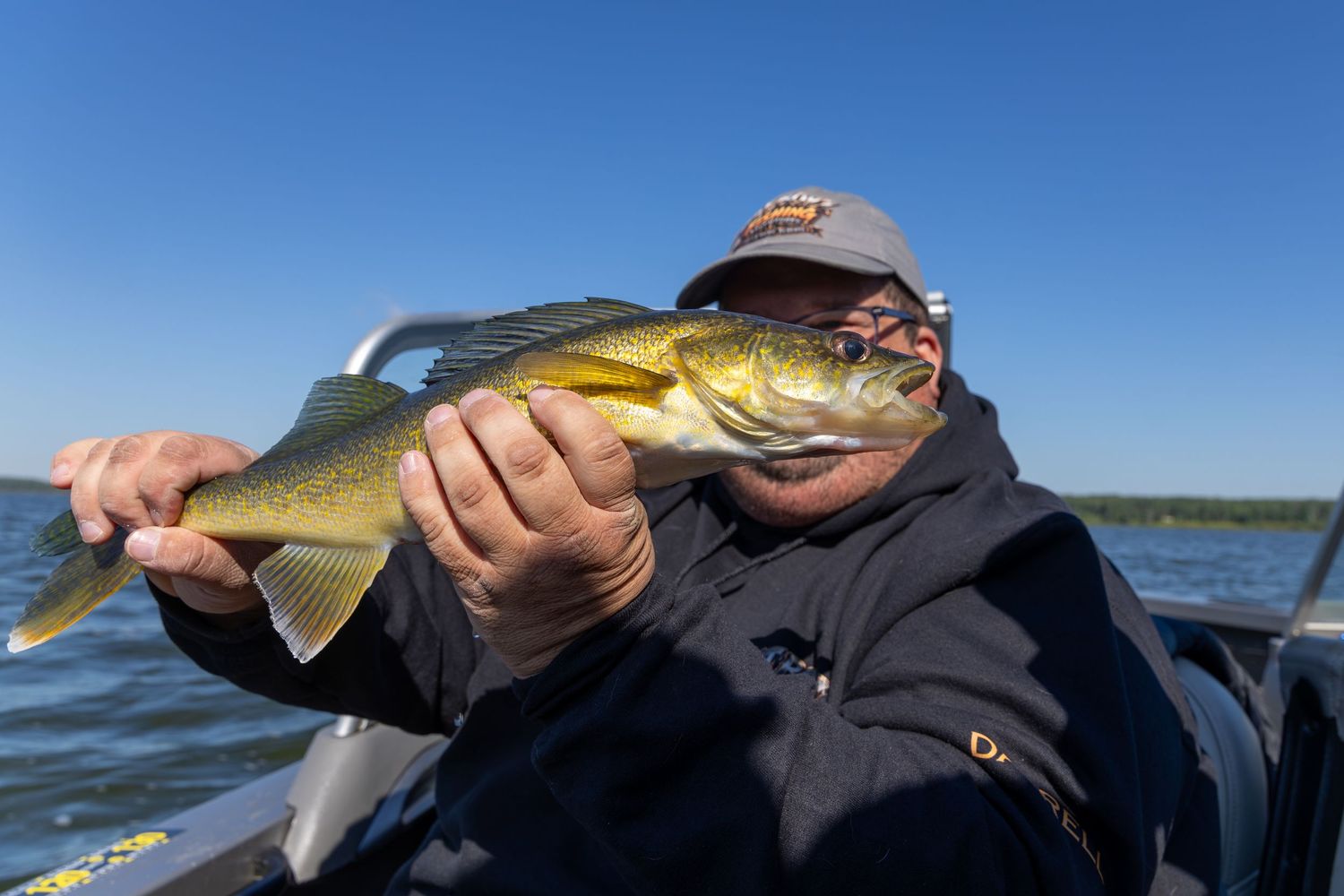 Angler holding a large walleye fish on a boat with blue sky and calm lake water in the background.