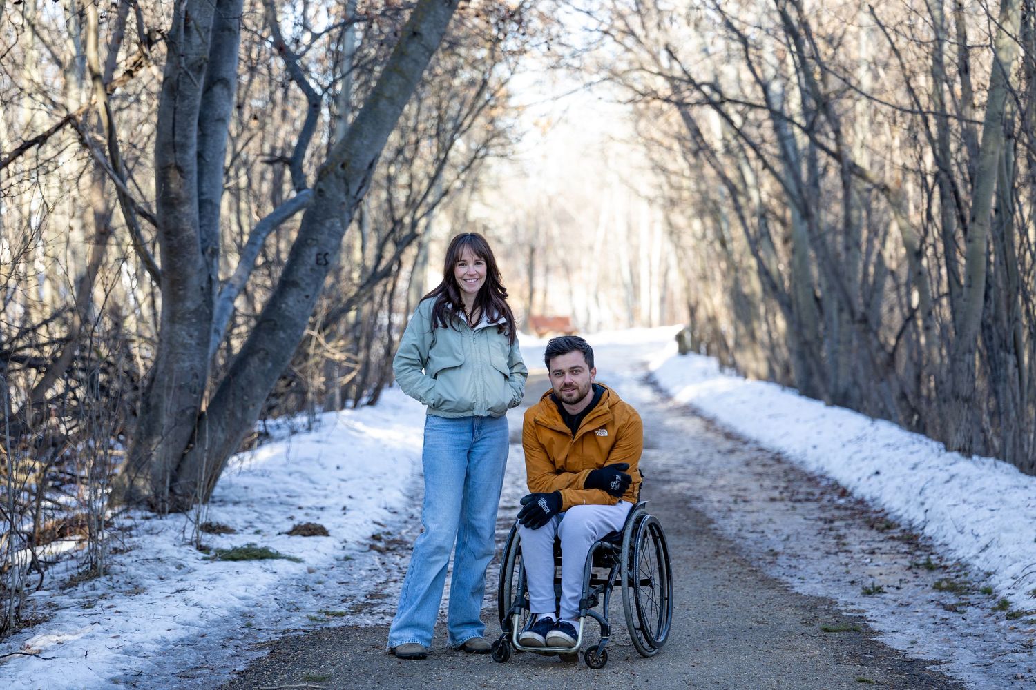 A woman stands beside a man in a wheelchair on a snowy forest path in winter, both smiling outdoors.