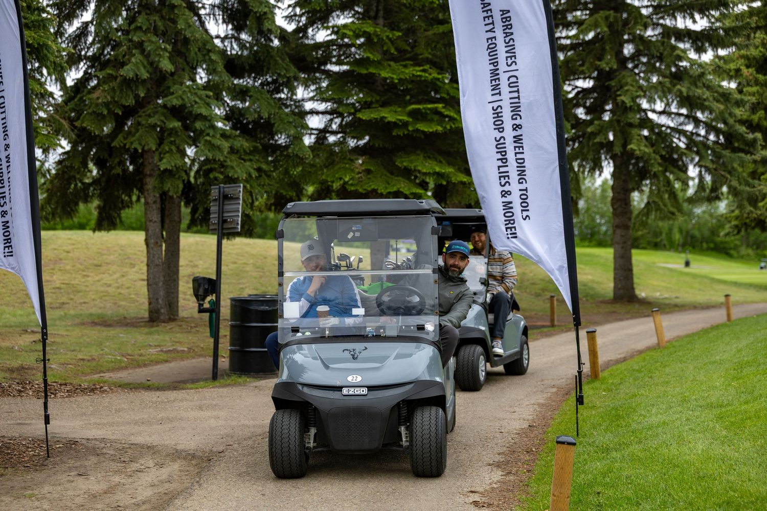 Two golf carts driving along a tree-lined path at a golf course charity event with sponsor banners.
