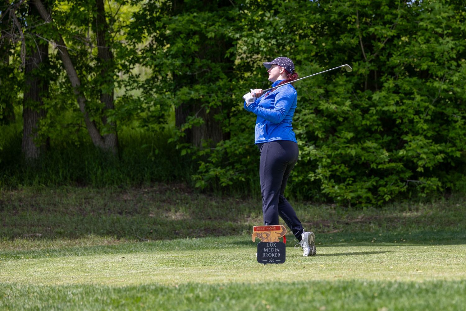 Golfer in blue jacket swinging club on lush green fairway surrounded by trees on a sunny day.