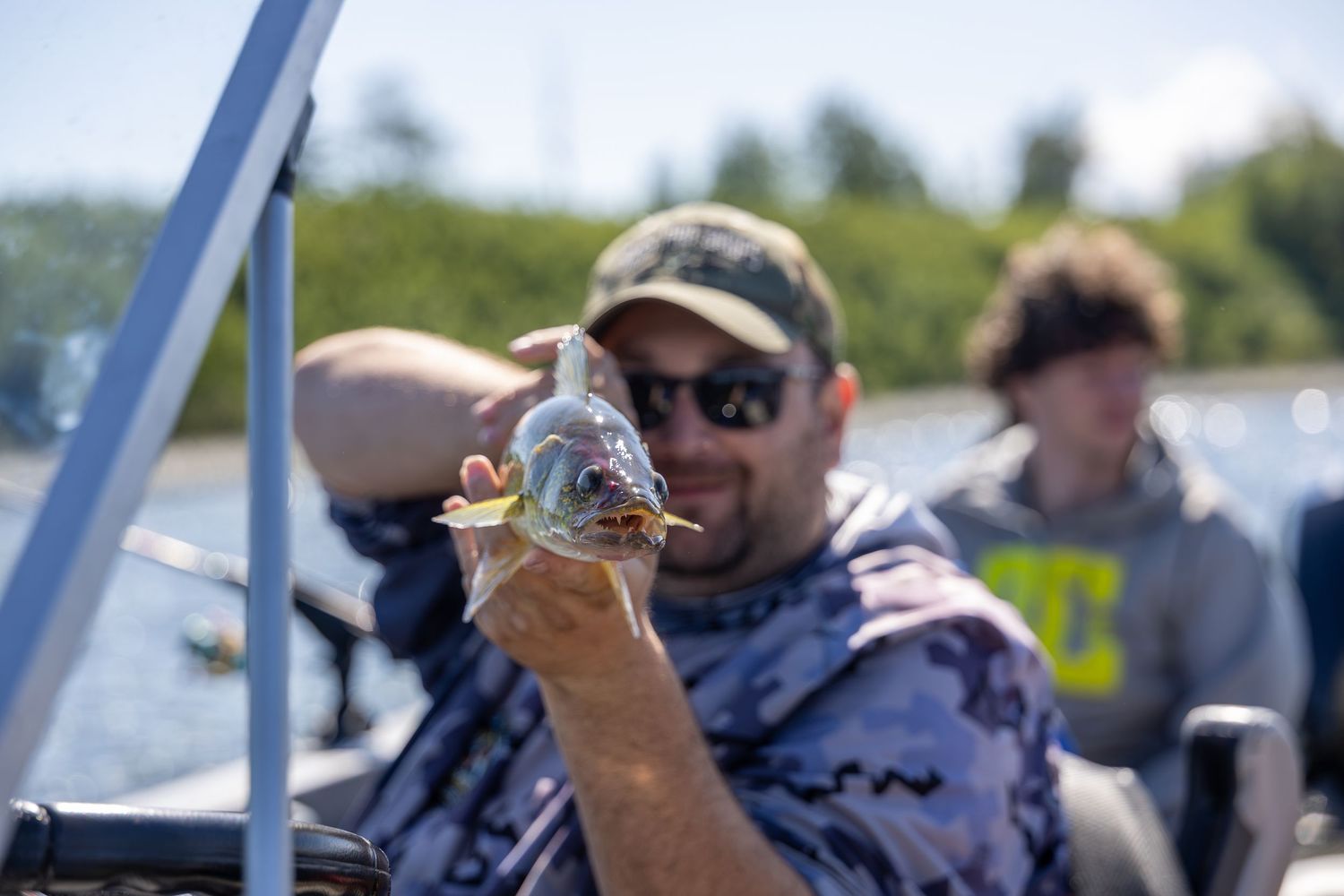 Man on a boat proudly holds up a small fish he caught while fishing outdoors on a sunny day.
