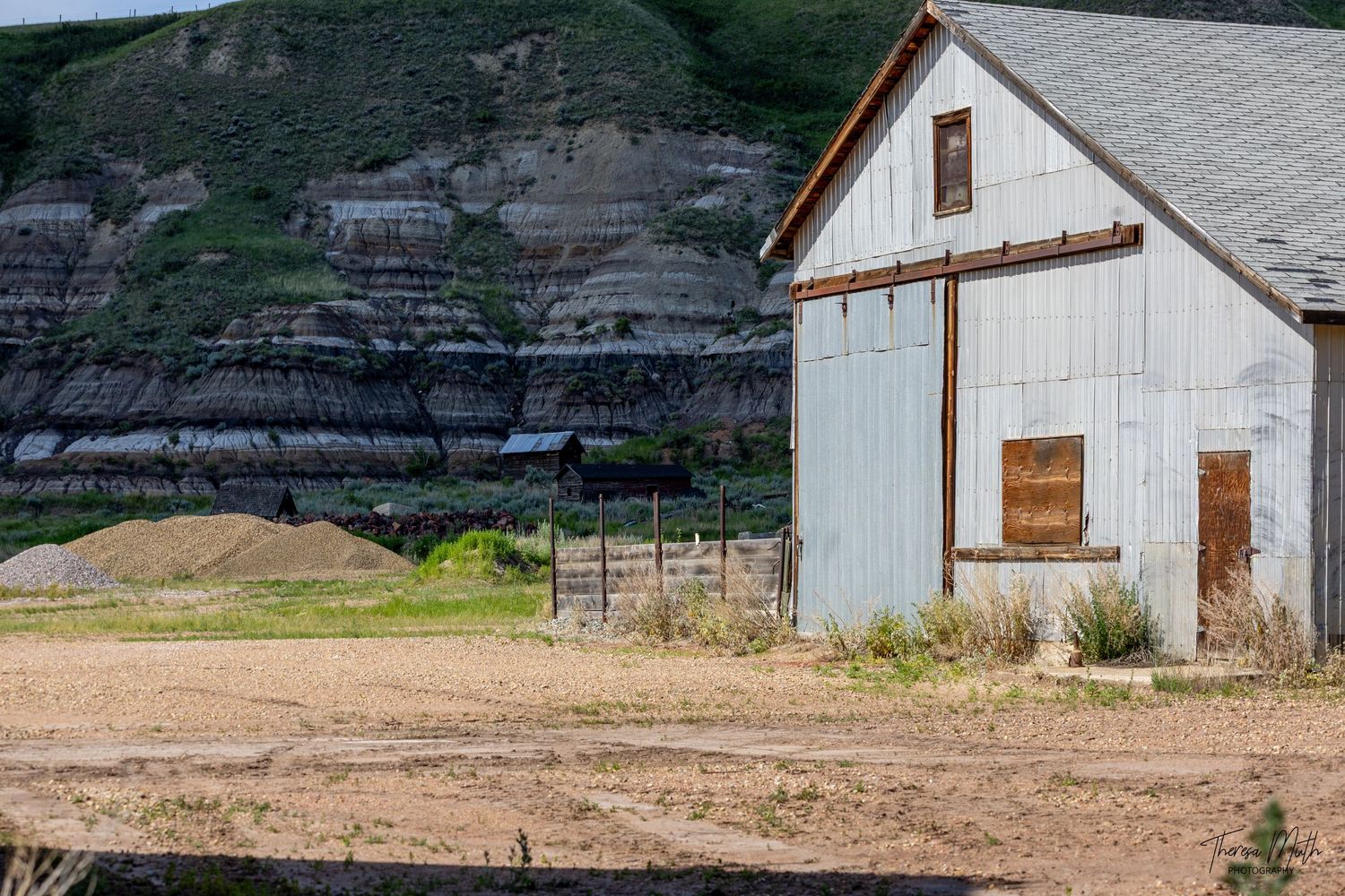 Rustic white barn beside dramatic layered rock cliffs in a rural landscape with dry dirt ground and green hillside.