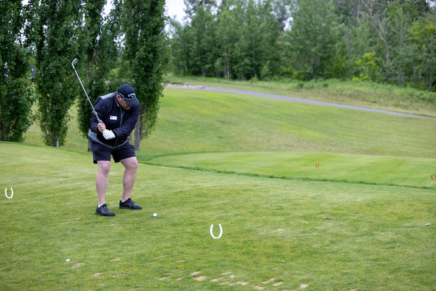 Golfer mid-swing on a lush green golf course, wearing black attire, with trees in the background.