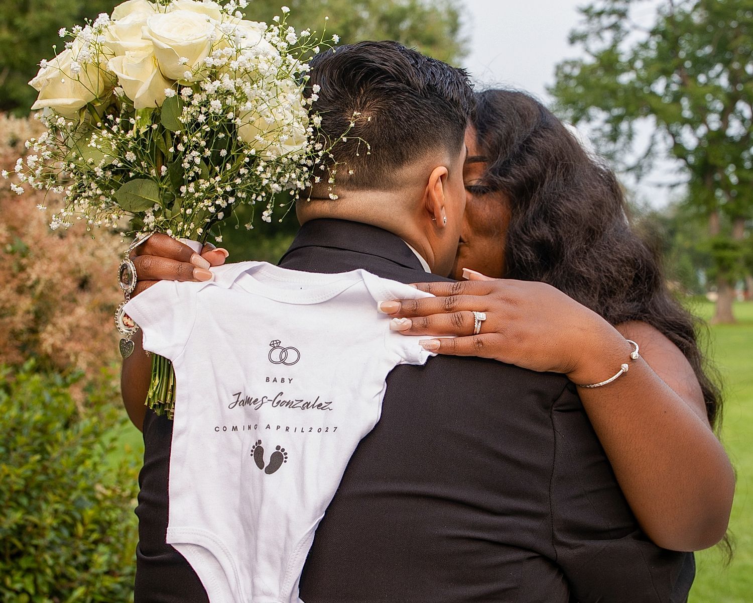 Couple embracing at wedding holding baby onesie and white rose bouquet, announcing pregnancy outdoors.