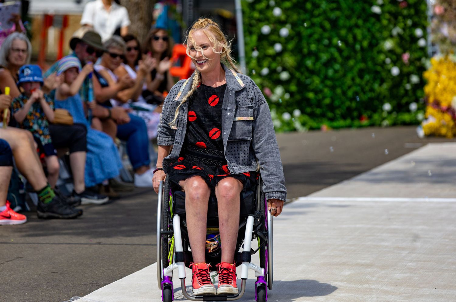 Young blonde woman in a wheelchair wearing a denim jacket and floral dress on an outdoor fashion runway.