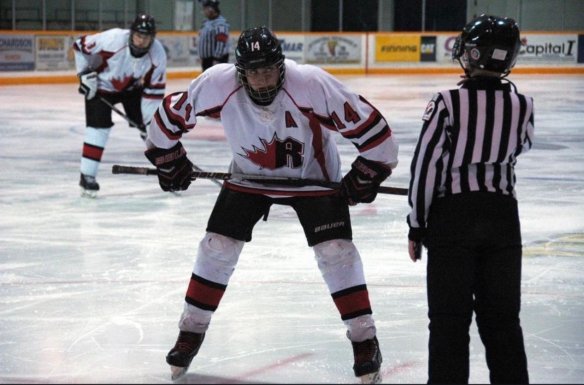Canadian hockey player in white jersey stands ready on ice rink during game with referee nearby.