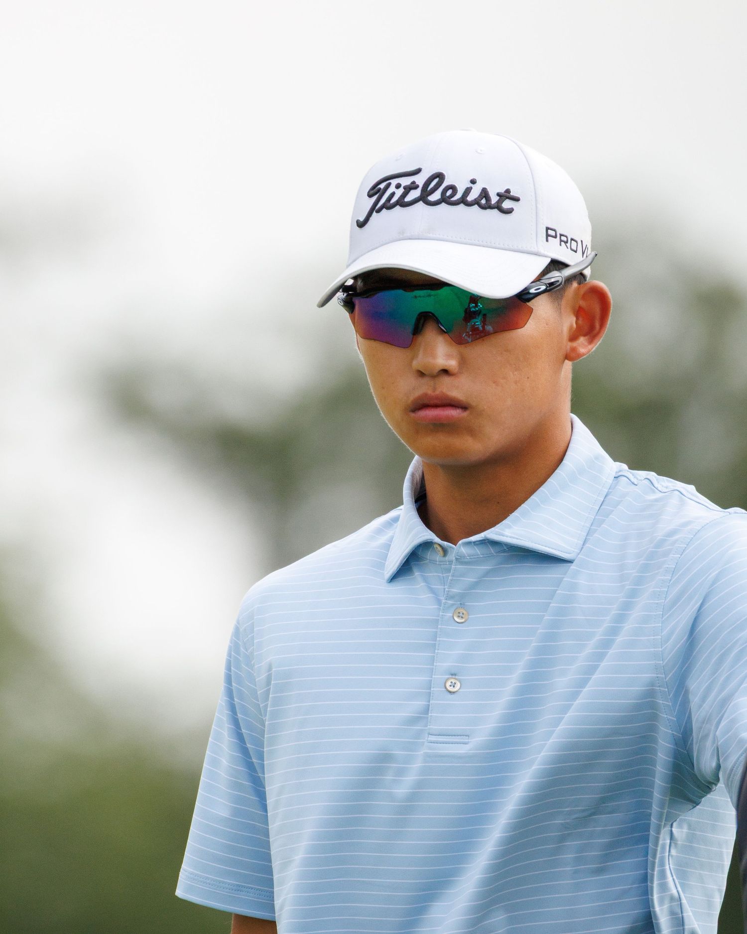 Young male golfer wearing a Titleist cap and blue polo shirt with reflective sunglasses on a golf course.