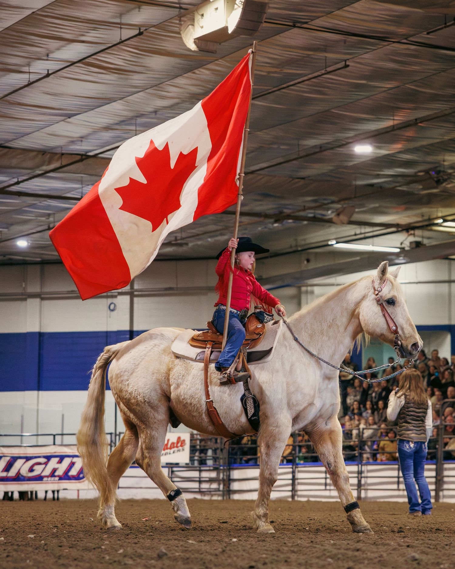 Rider on white horse carrying Canadian flag at indoor rodeo arena.