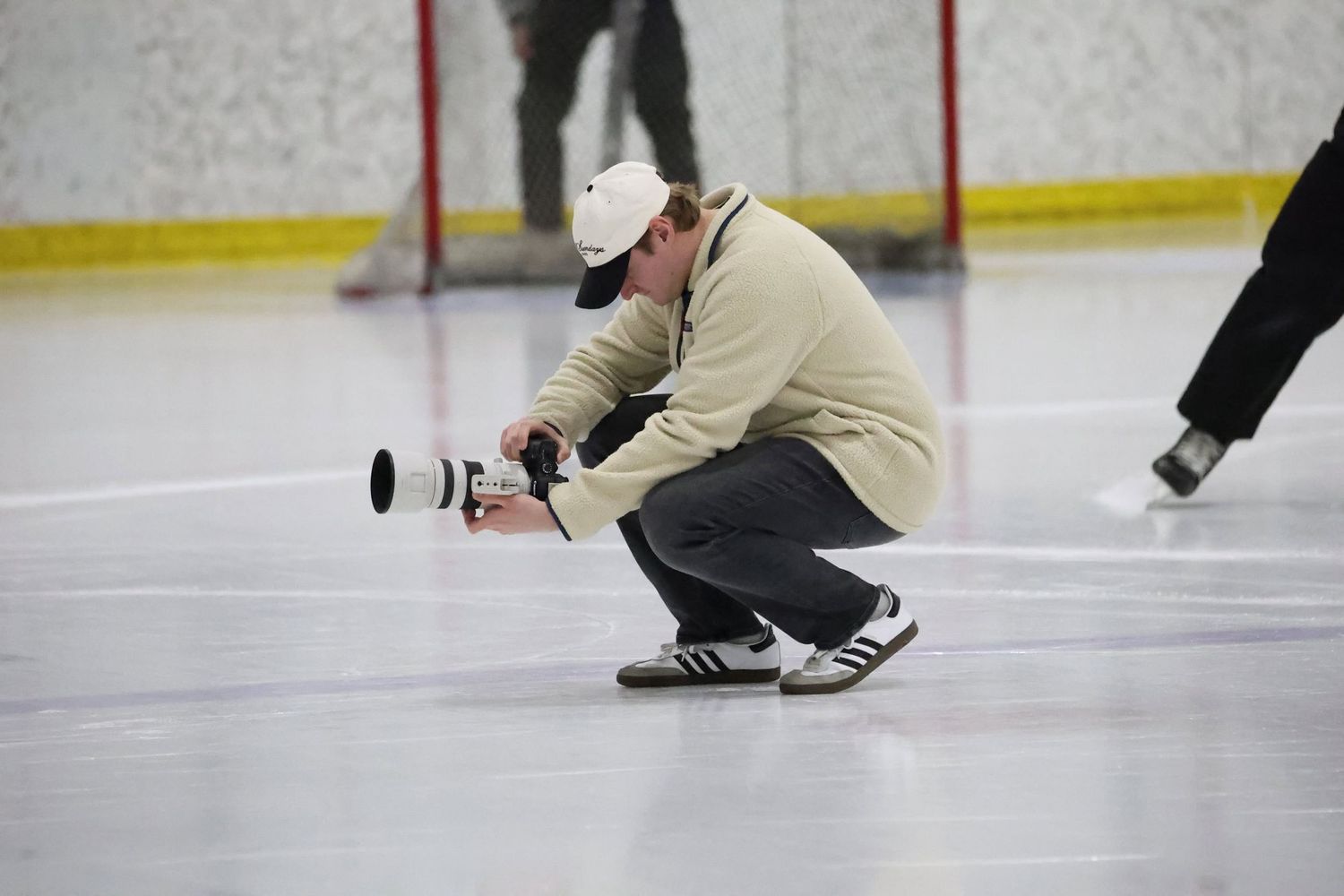Photographer crouching on ice rink with large camera lens capturing hockey game action at indoor arena.