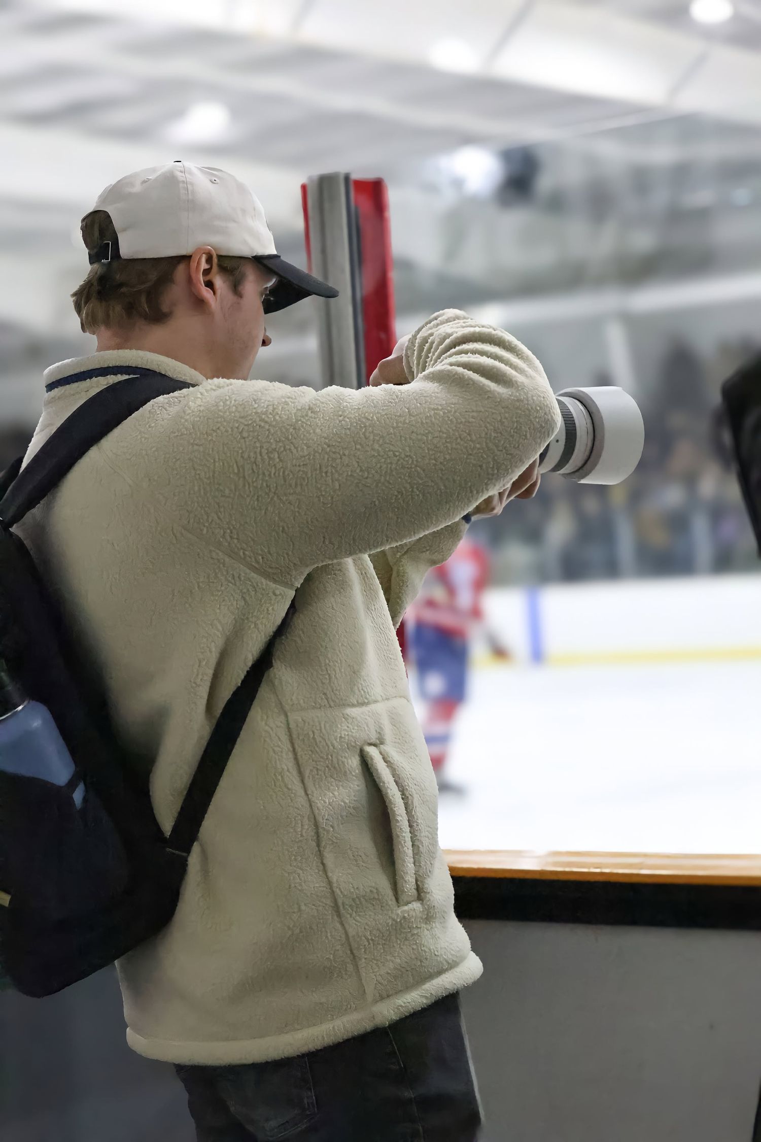 Photographer with telephoto lens and backpack captures hockey game action at indoor ice rink.