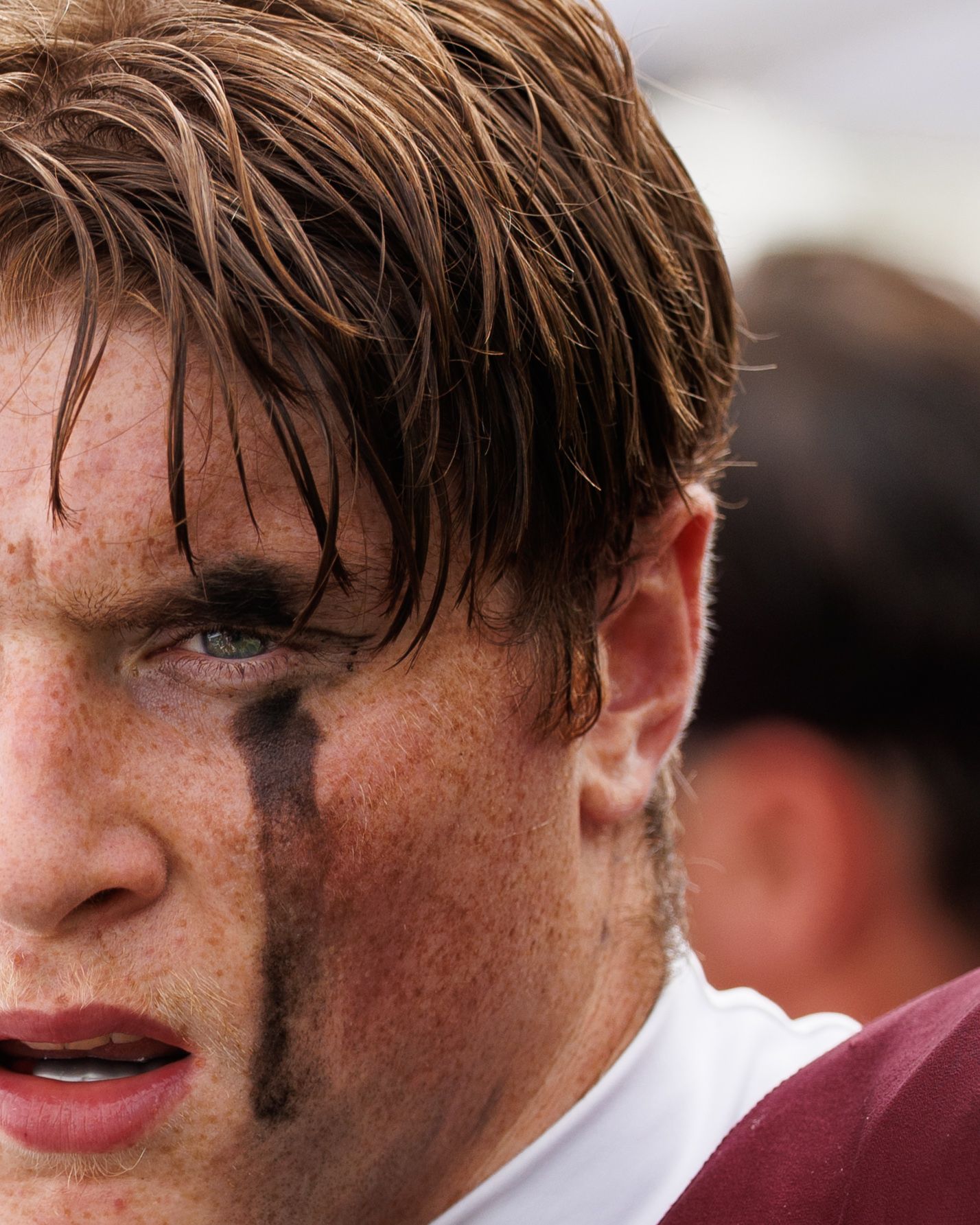 Close-up of a sweaty male athlete with eye black on his cheek, wet hair, and freckles during a sports game.