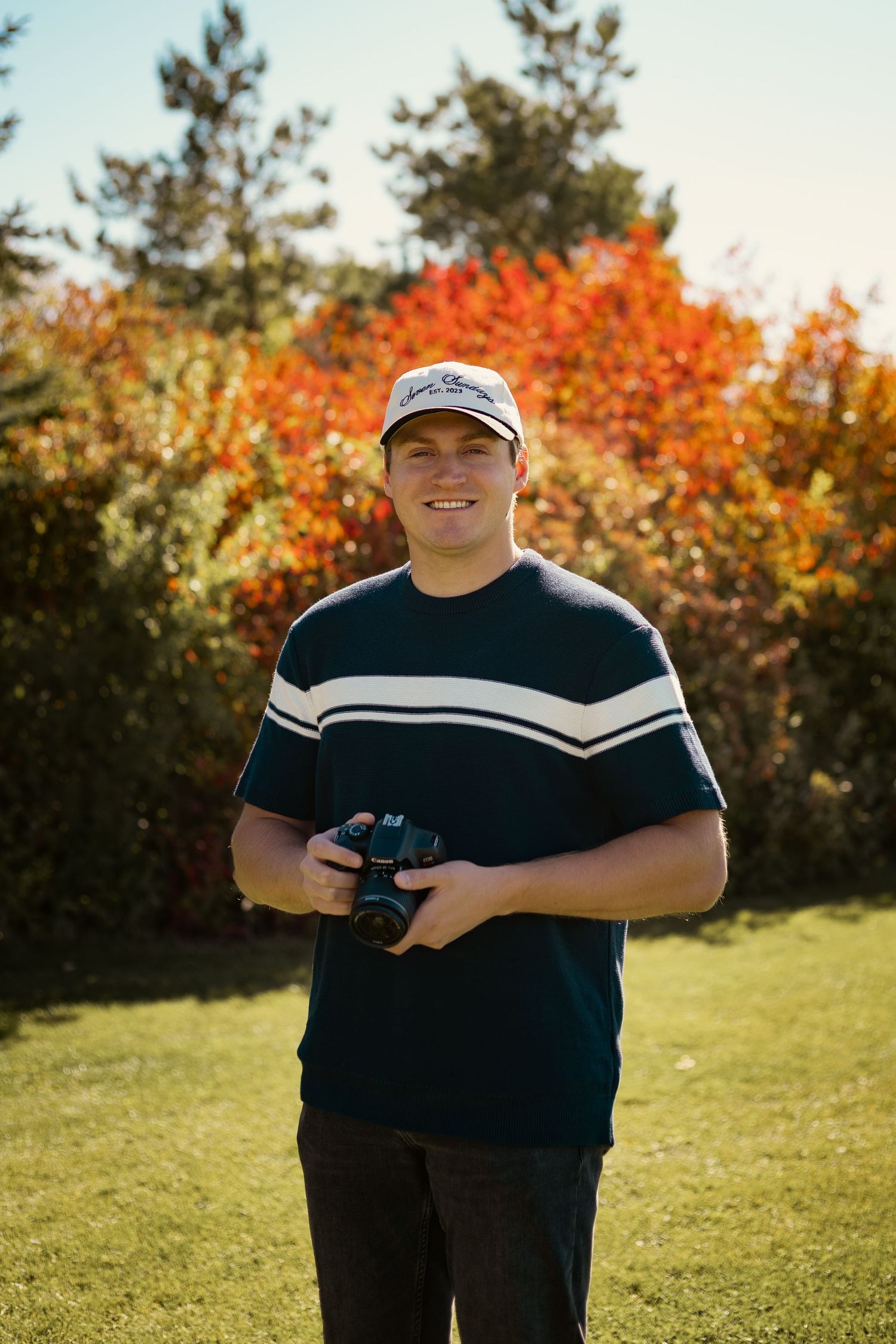 Young man smiling outdoors holding a camera, wearing a cap and navy striped shirt, with vibrant autumn foliage behind him.