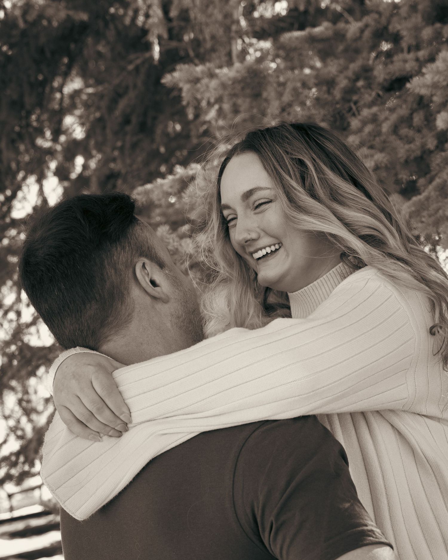 Black and white photo of a laughing woman hugging a man outdoors under trees in a romantic embrace.