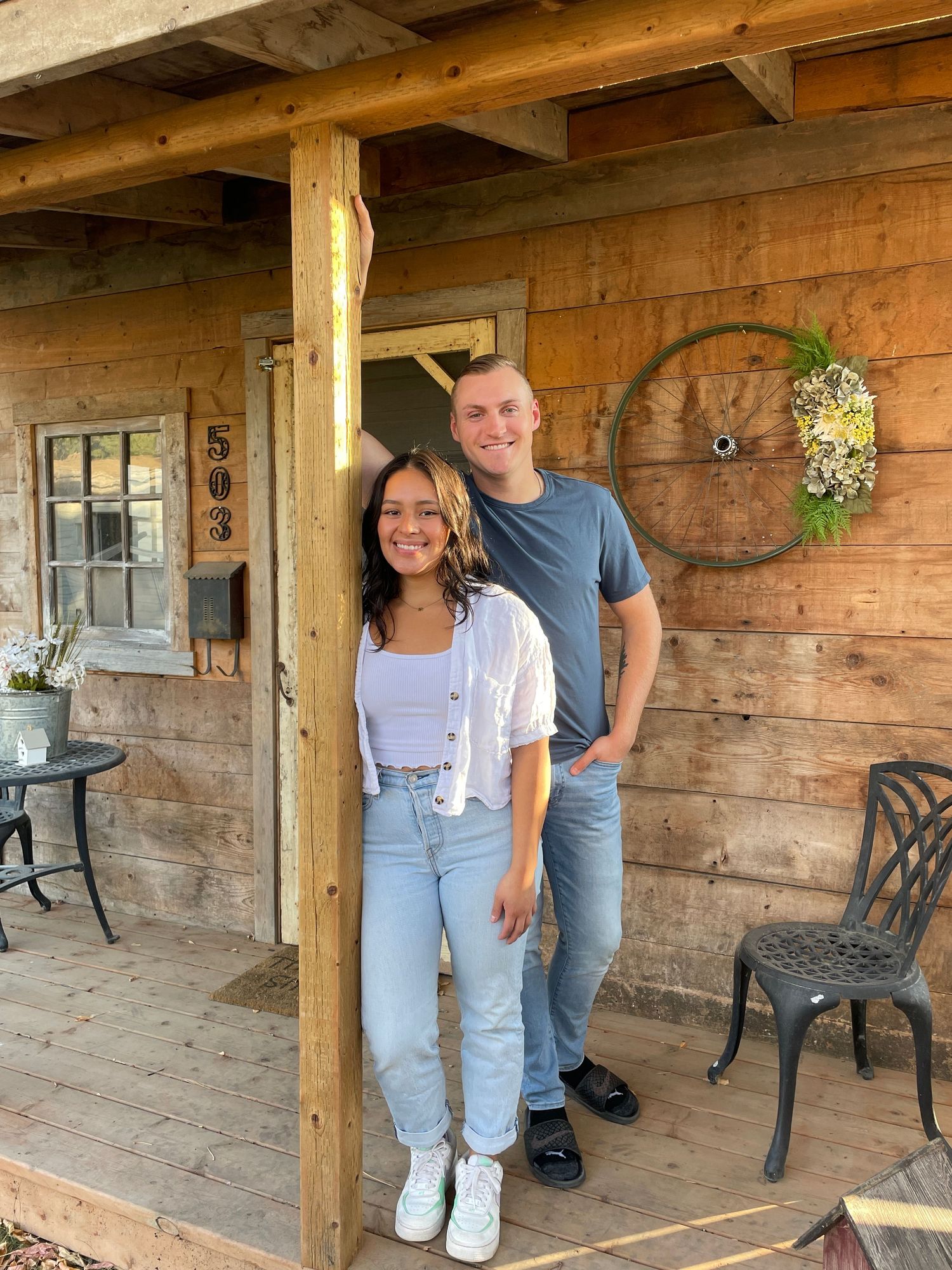 A smiling couple poses on a rustic wooden cabin porch with a decorative wheel wreath and house number 503.