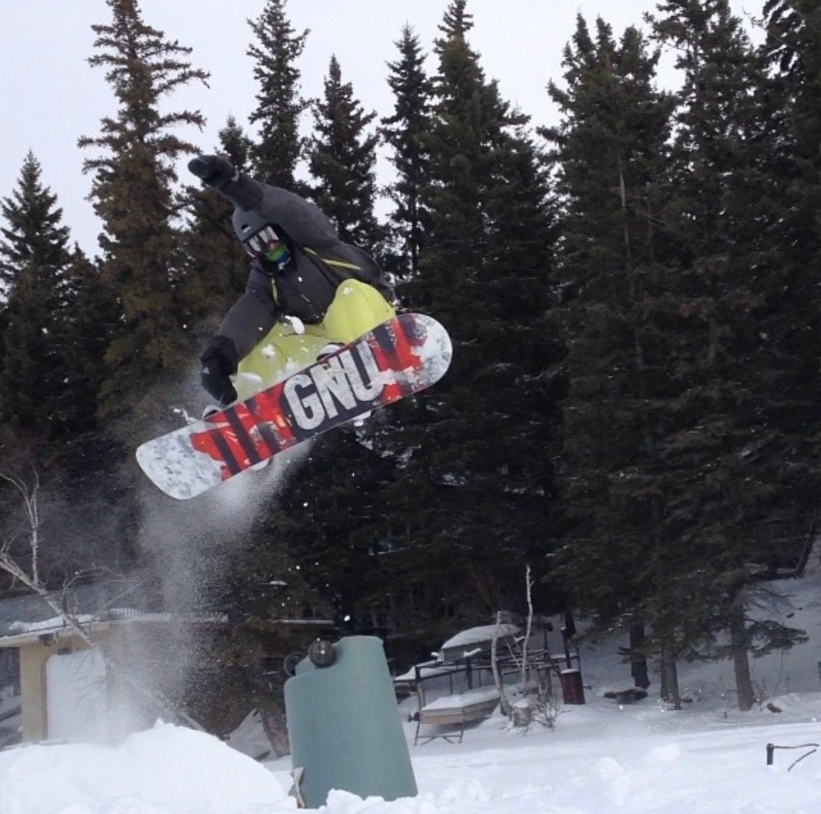 Snowboarder performing a high jump trick on a GNU snowboard amid snowy pine trees at a winter resort.