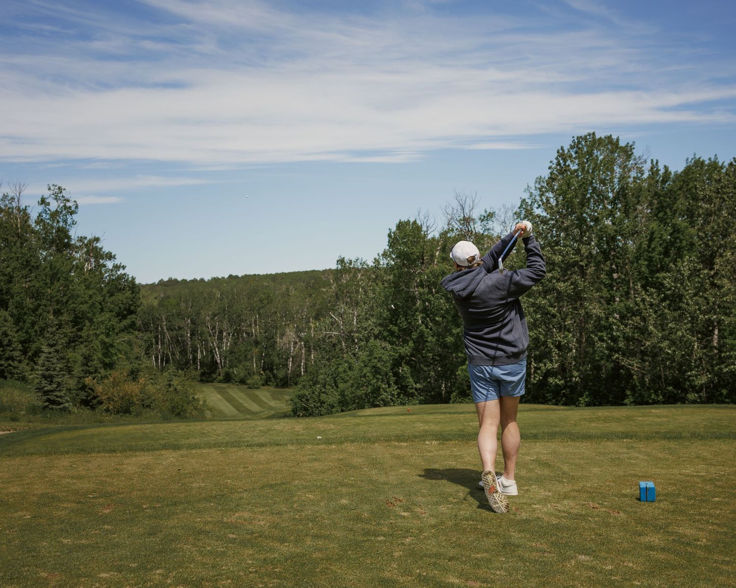 Golfer completing a swing on a lush green golf course surrounded by trees under a blue sky.