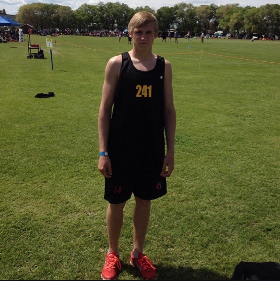 Young male athlete wearing black tank top with number 241, standing on green sports field in red sneakers.