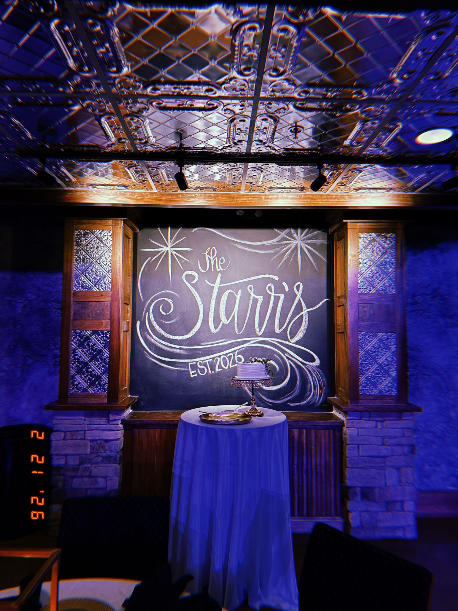 A series of illuminated chalkboard signs with decorative text and blue curtains in a dimly lit brick wall venue setting.