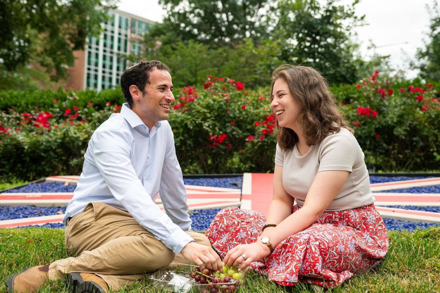Couple relaxing on grass in a garden with red flowers and blue borders in the background.