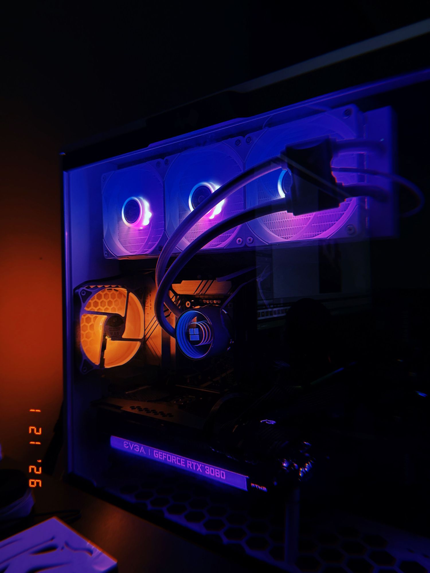 Gaming setup with orange and purple LED lighting illuminating multiple monitors and gaming peripherals in a dark room.