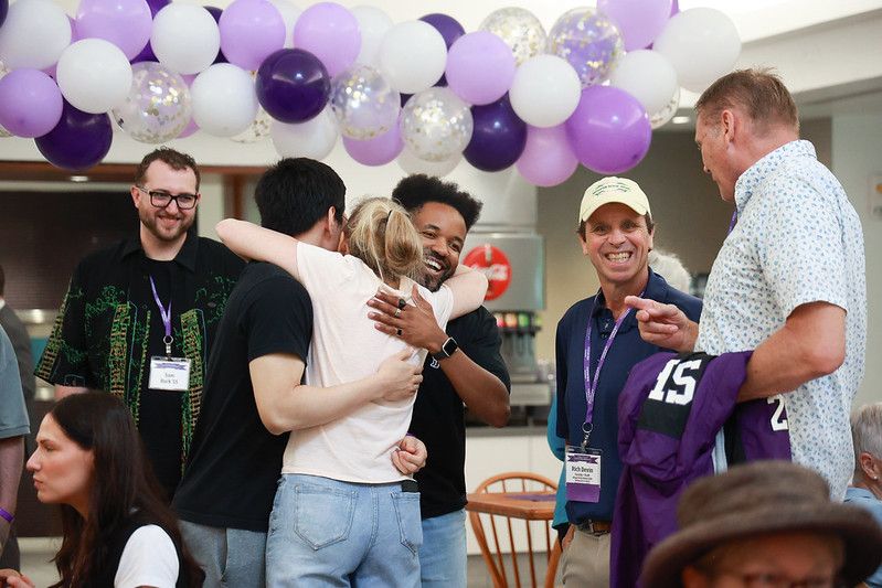 Group of people socializing at an event with purple and white balloon decorations in a room.