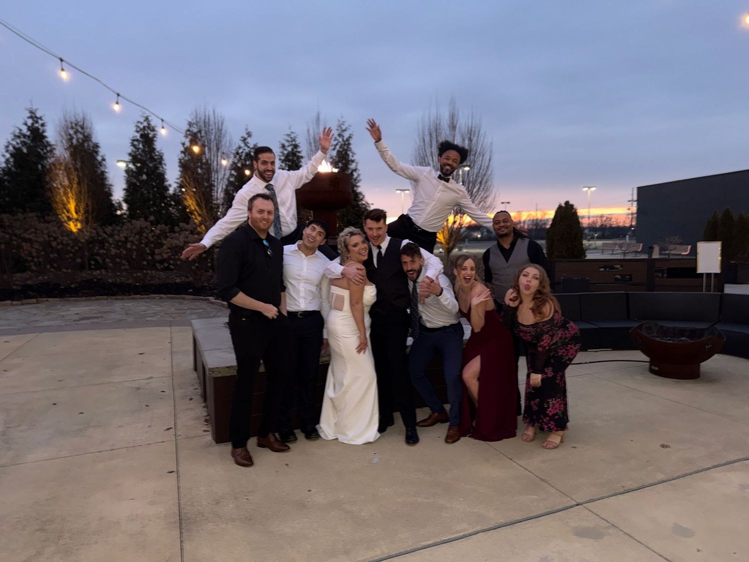 Wedding party celebrates with joyful poses on an outdoor patio at dusk with string lights overhead.