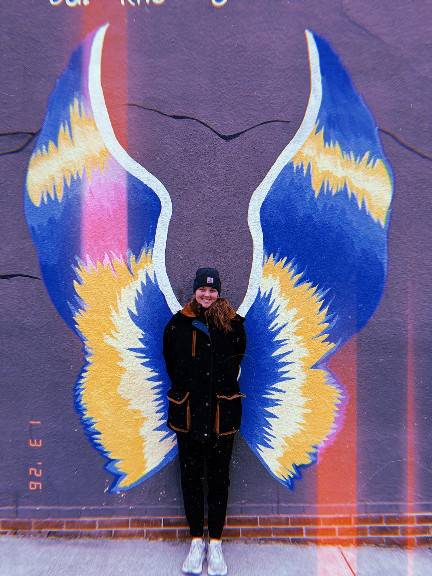 A person poses in front of vibrant blue and yellow angel wings street art mural painted on a purple wall.