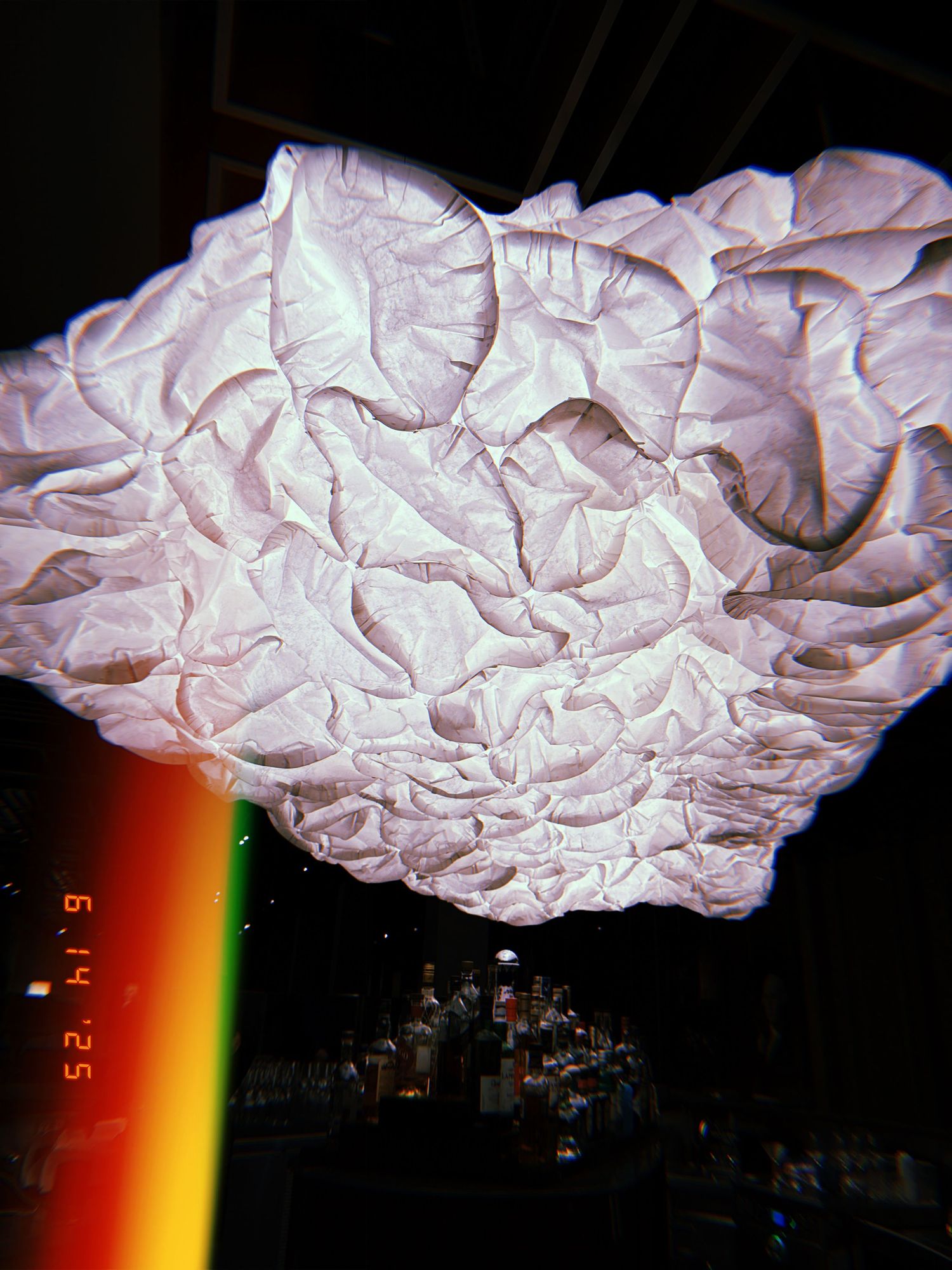 A pink crumpled paper installation piece hangs against a dark background with a rainbow light streak in the foreground.