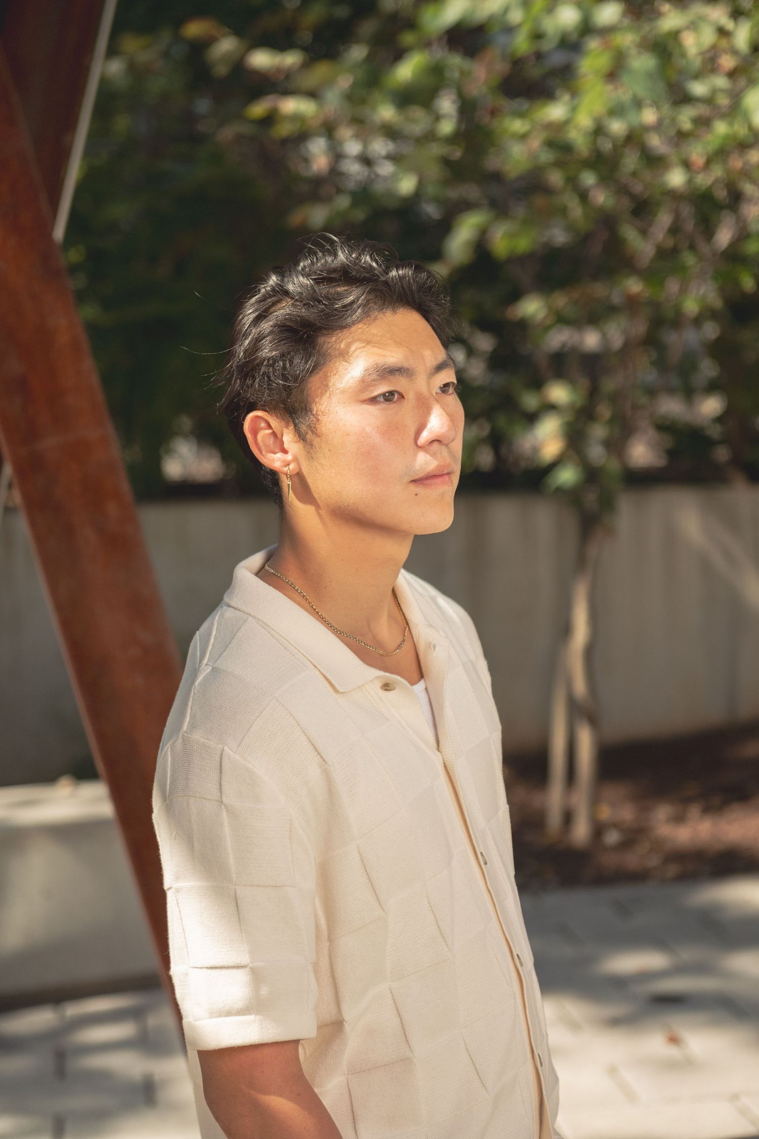 A person in a beige short-sleeved button-up shirt poses outdoors against a sunlit background with trees.