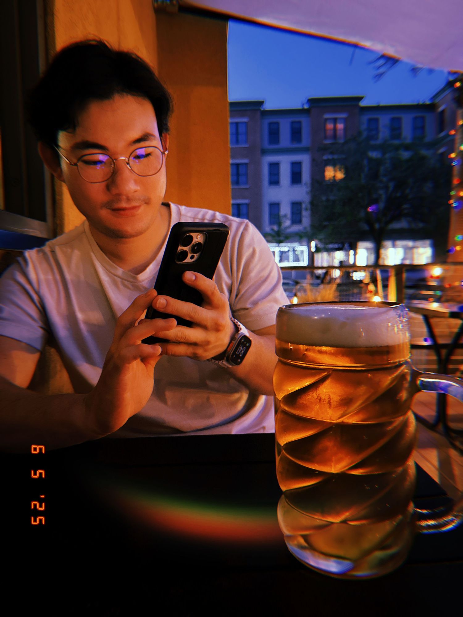 Person checking their phone while sitting at a restaurant with a uniquely designed beer glass and city buildings outside.