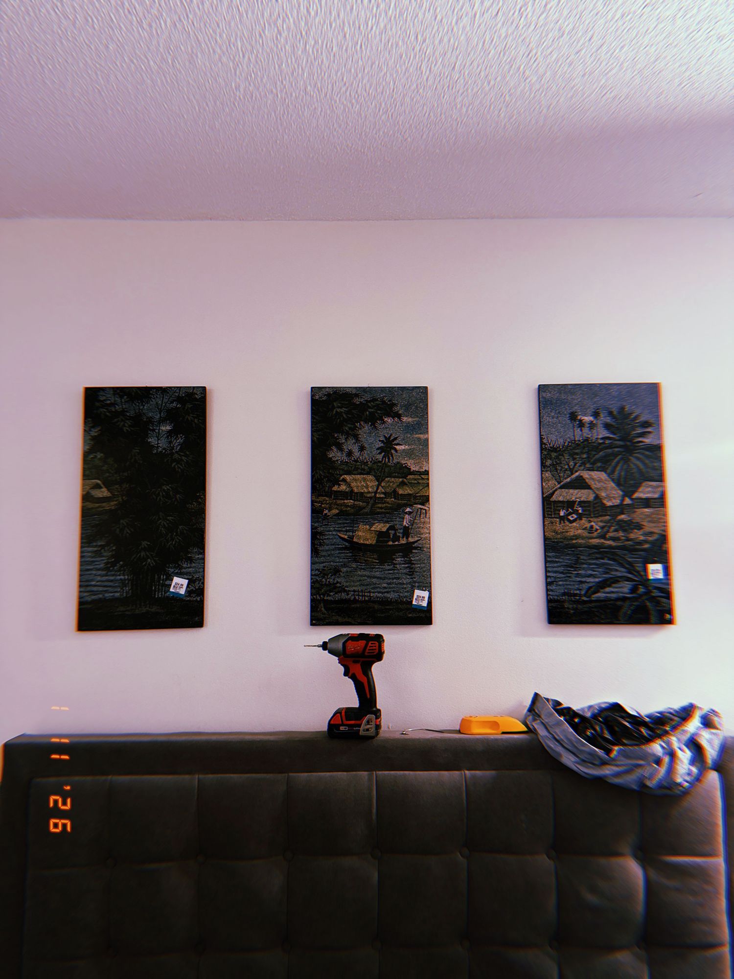 Three dark-framed landscape paintings hang on a white wall above a black leather couch with scattered pillows and blankets.