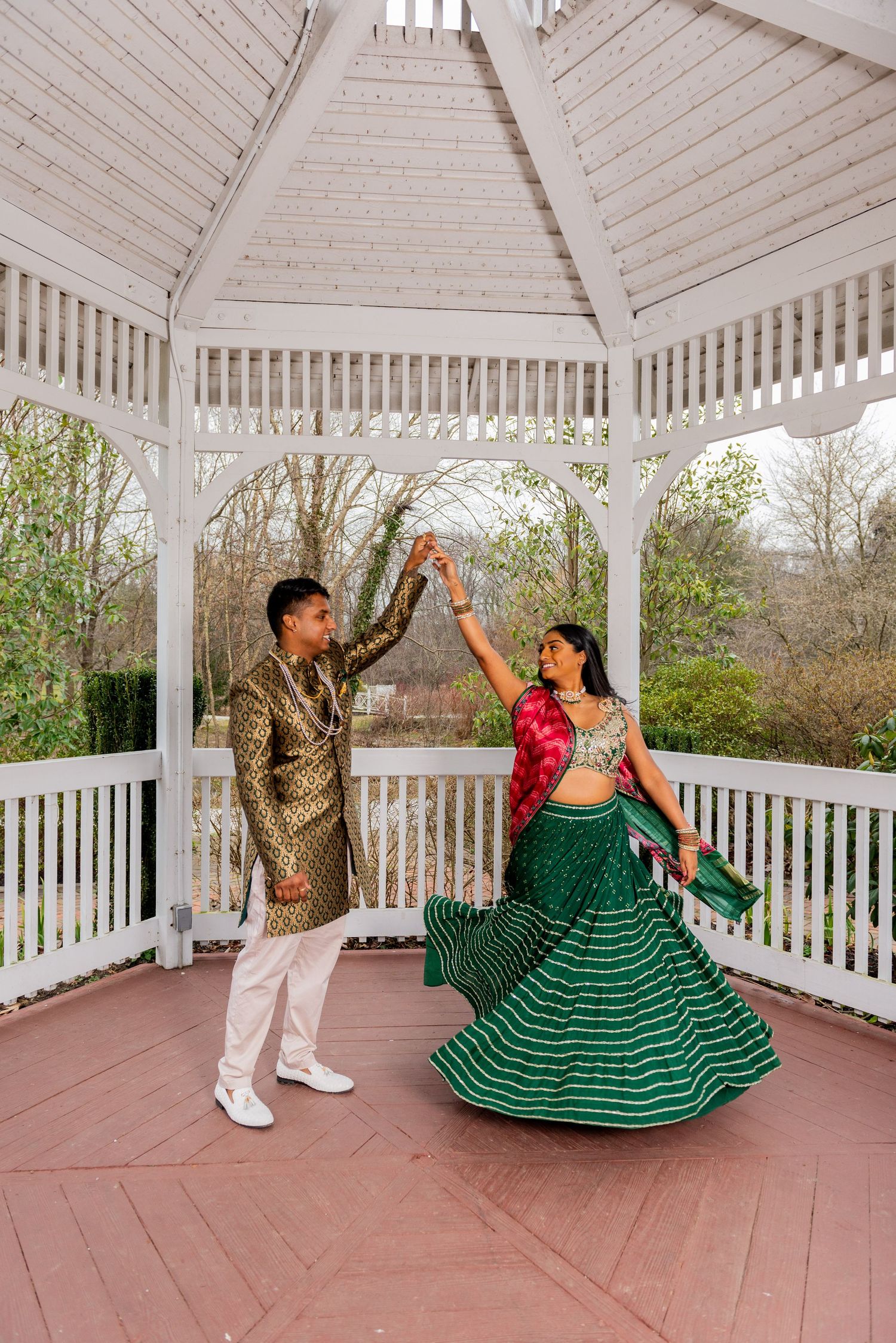 A couple in traditional Indian attire dances together in a white gazebo surrounded by greenery.