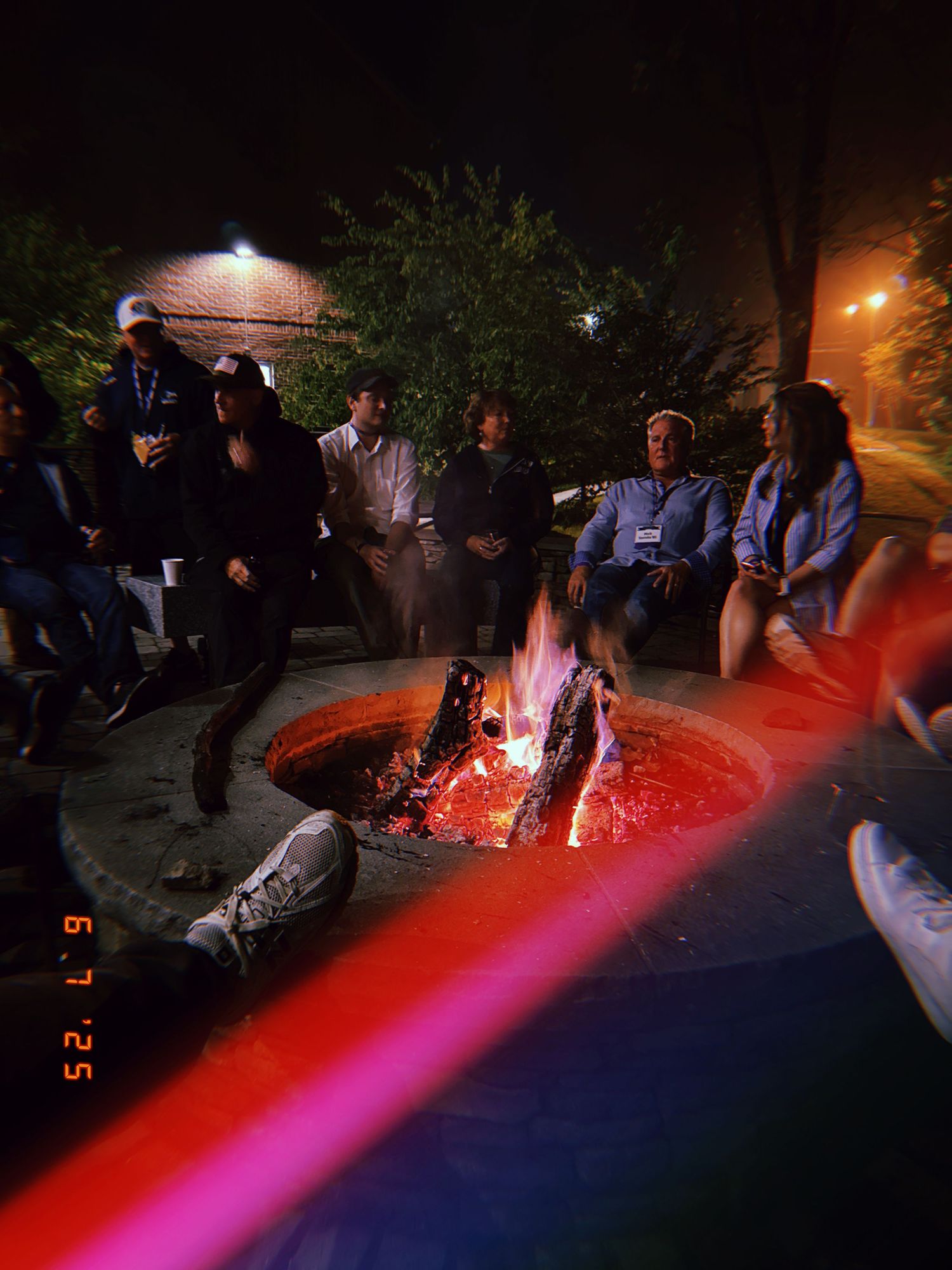 A group of people gathered around a glowing fire pit at night with outdoor lighting and trees in the background.