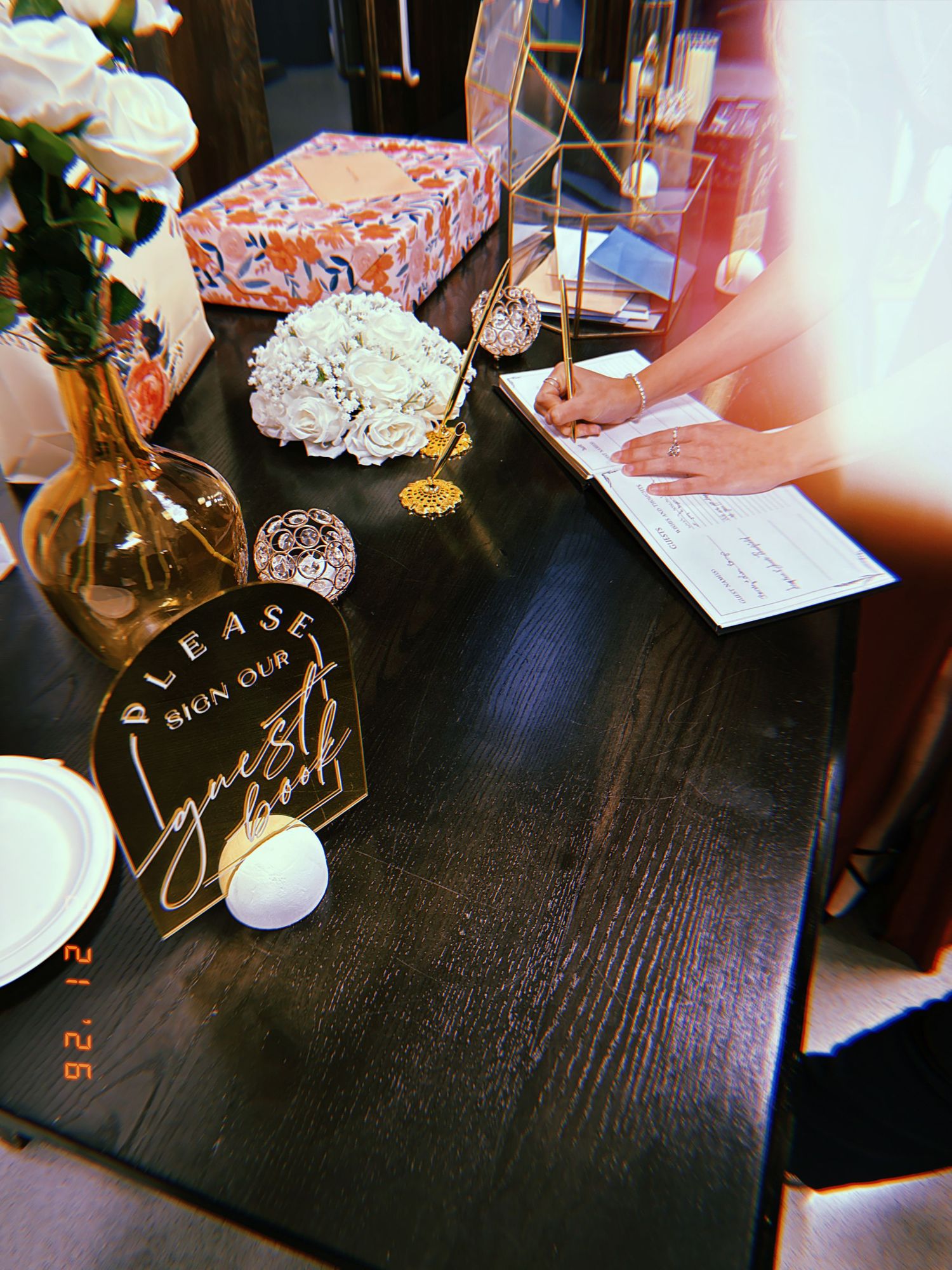 Someone writes at a dark wooden table decorated with party items, gifts, and floral arrangements in a warm ambient setting.