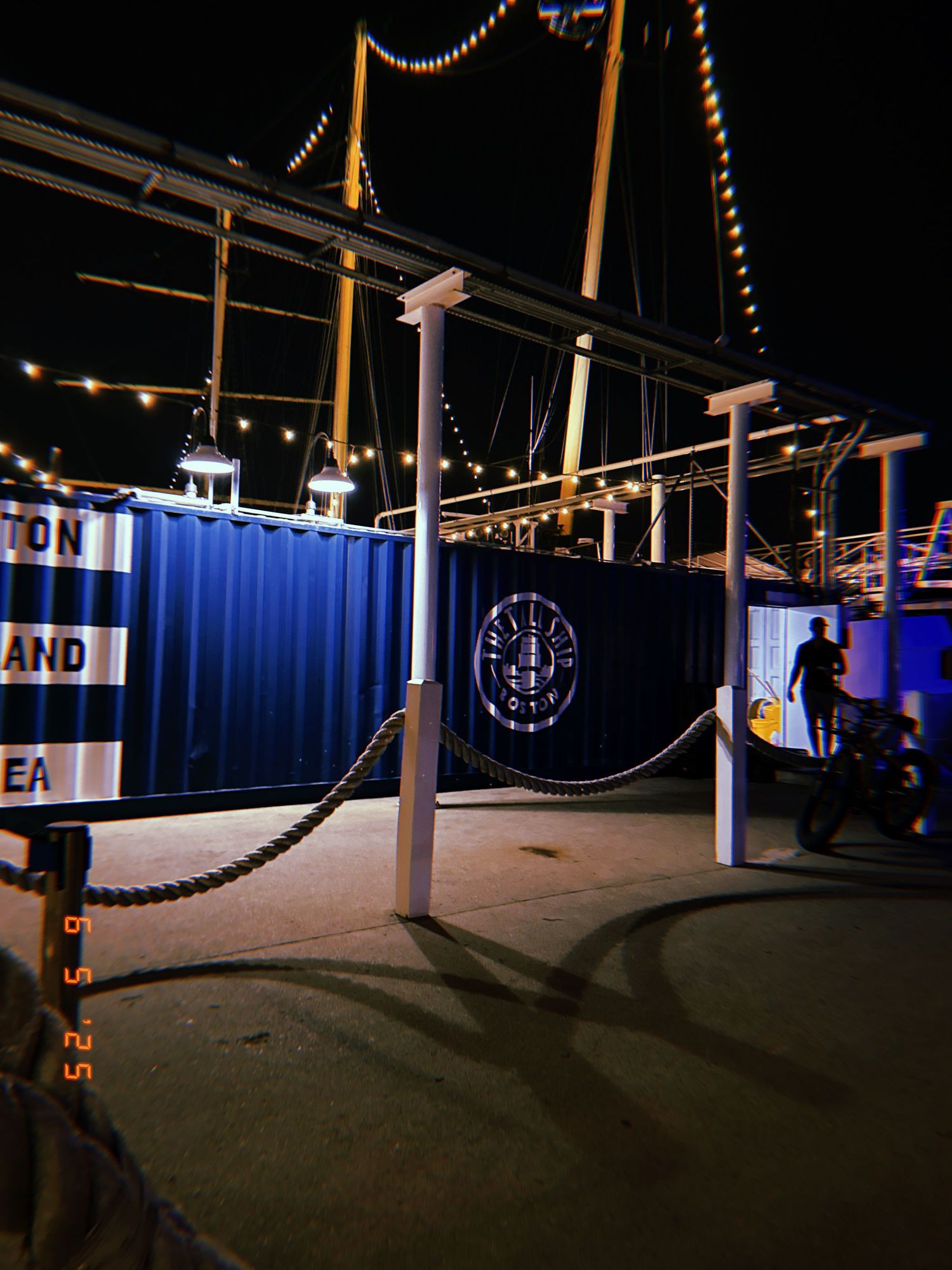 A nighttime shipping container venue with rope barriers and illuminated anchor logo glows under string lights.