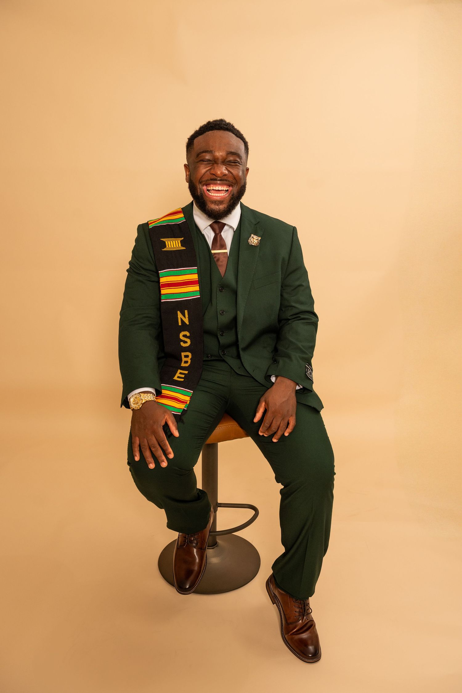 A graduate in a dark green academic regalia with colorful stoles poses in multiple celebratory positions against a beige backdrop.