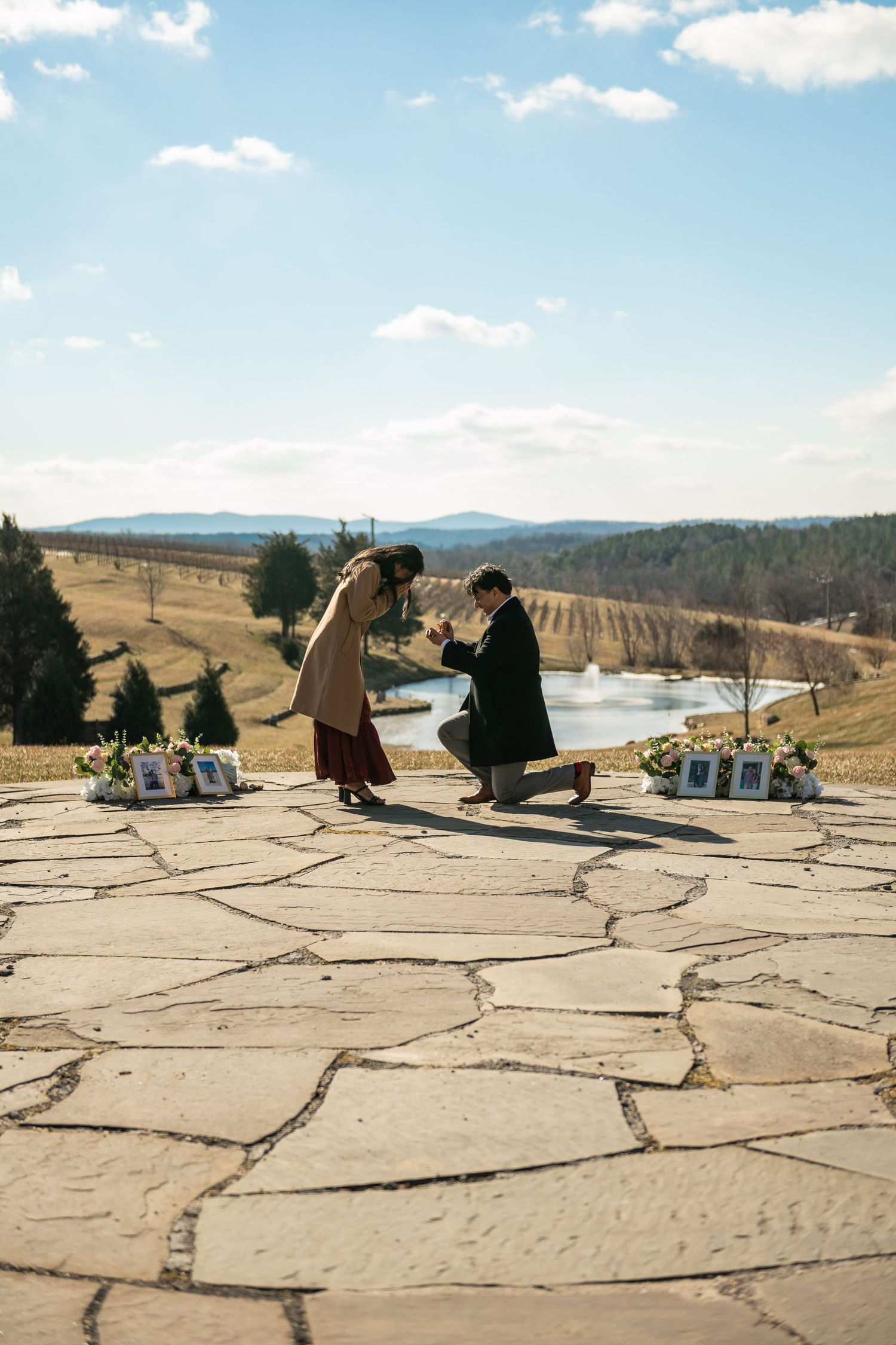A romantic proposal moment on a stone patio overlooking a scenic mountain valley with decorative floral arrangements.