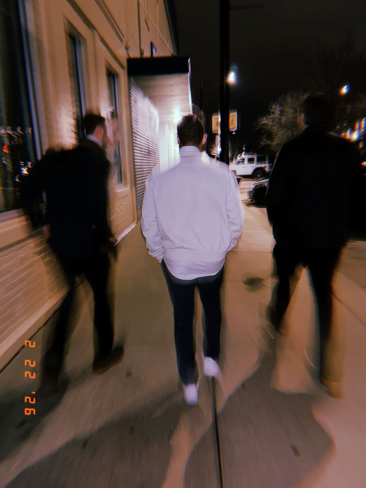 Three silhouetted figures walk along a dimly lit sidewalk at night with a blurred motion effect.