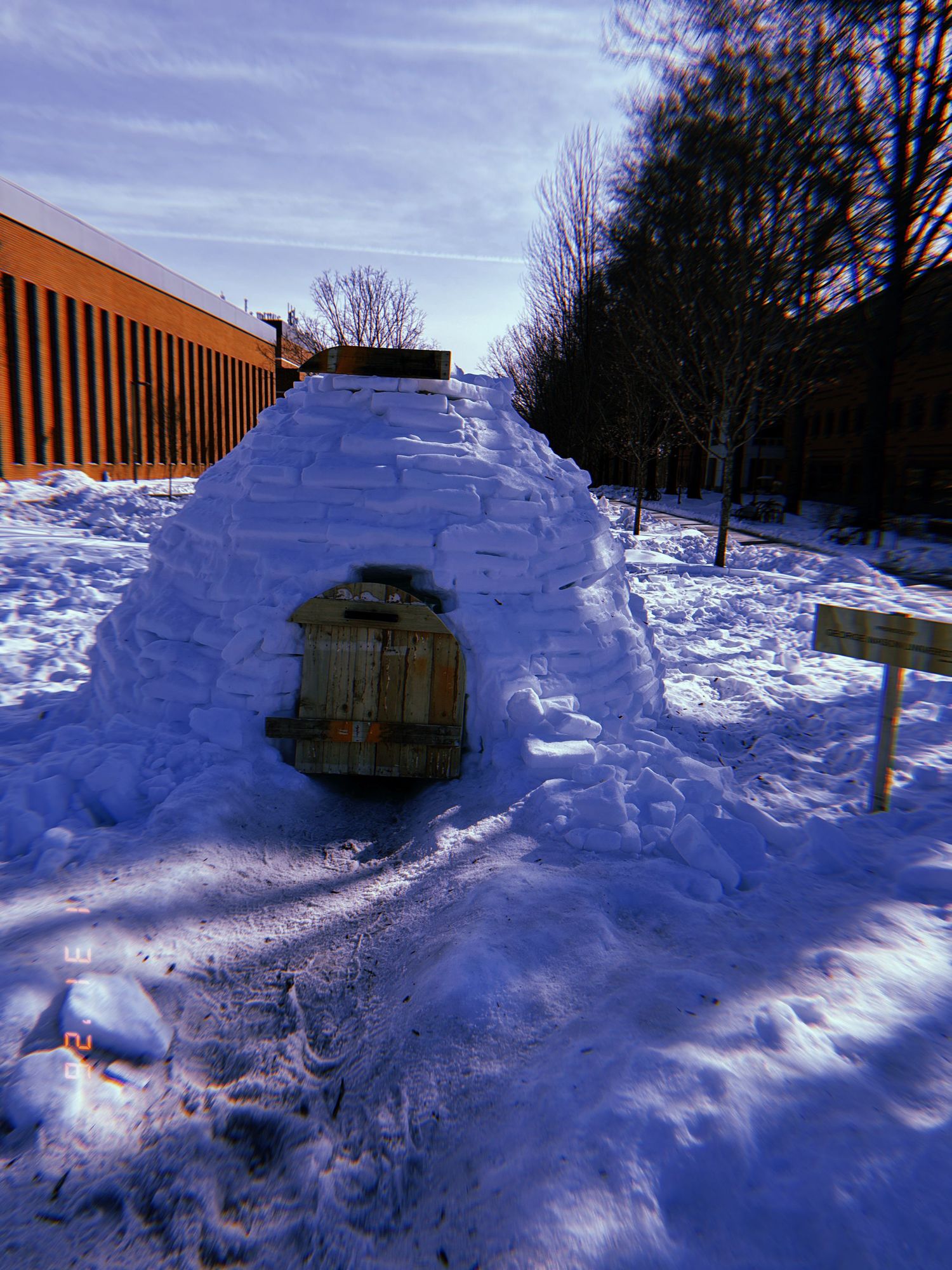 A snow igloo with a wooden door built in a winter landscape against an orange building.