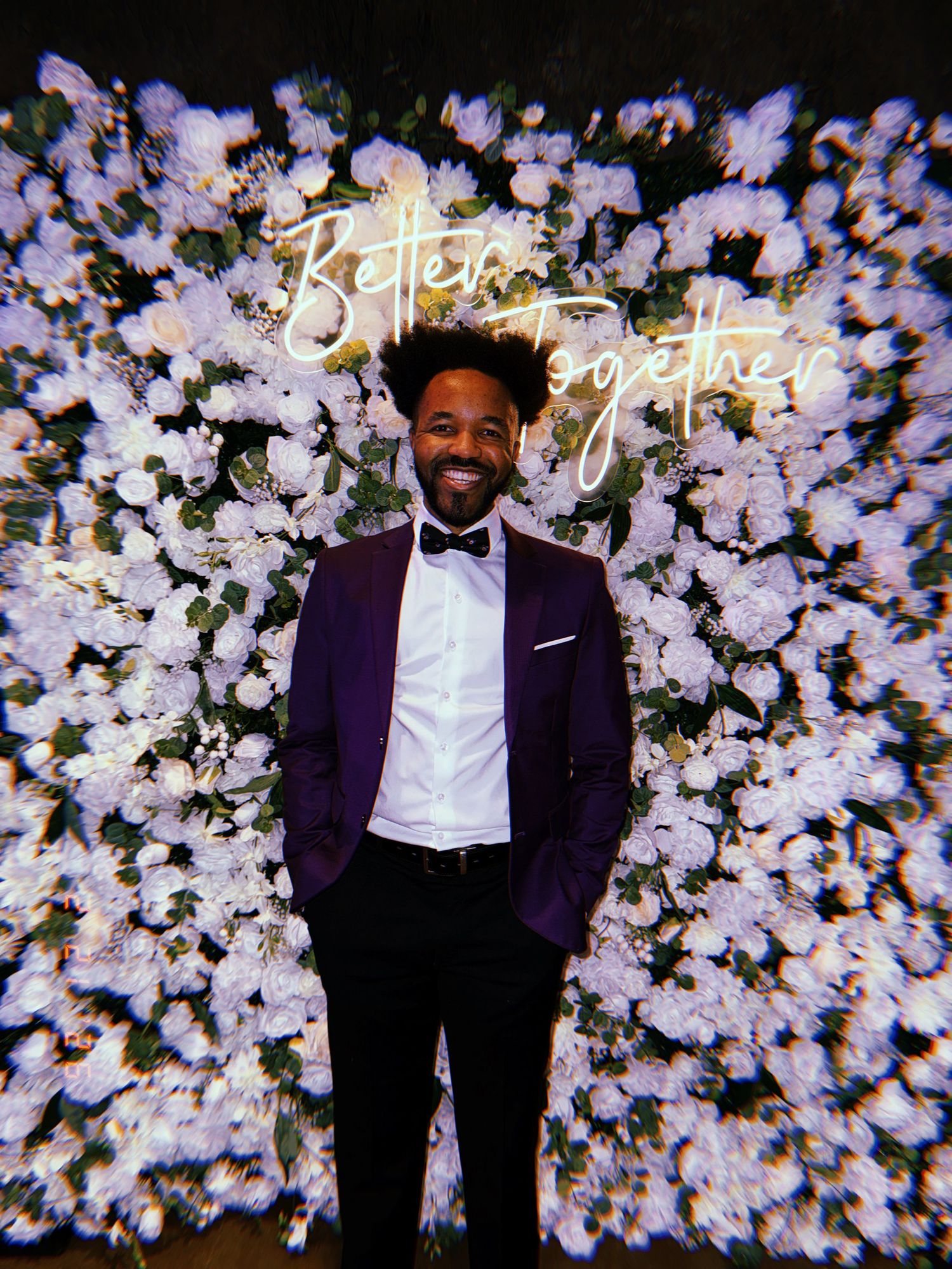 A person in a purple tuxedo jacket and bow tie stands in front of a wall of white flowers with illuminated text.