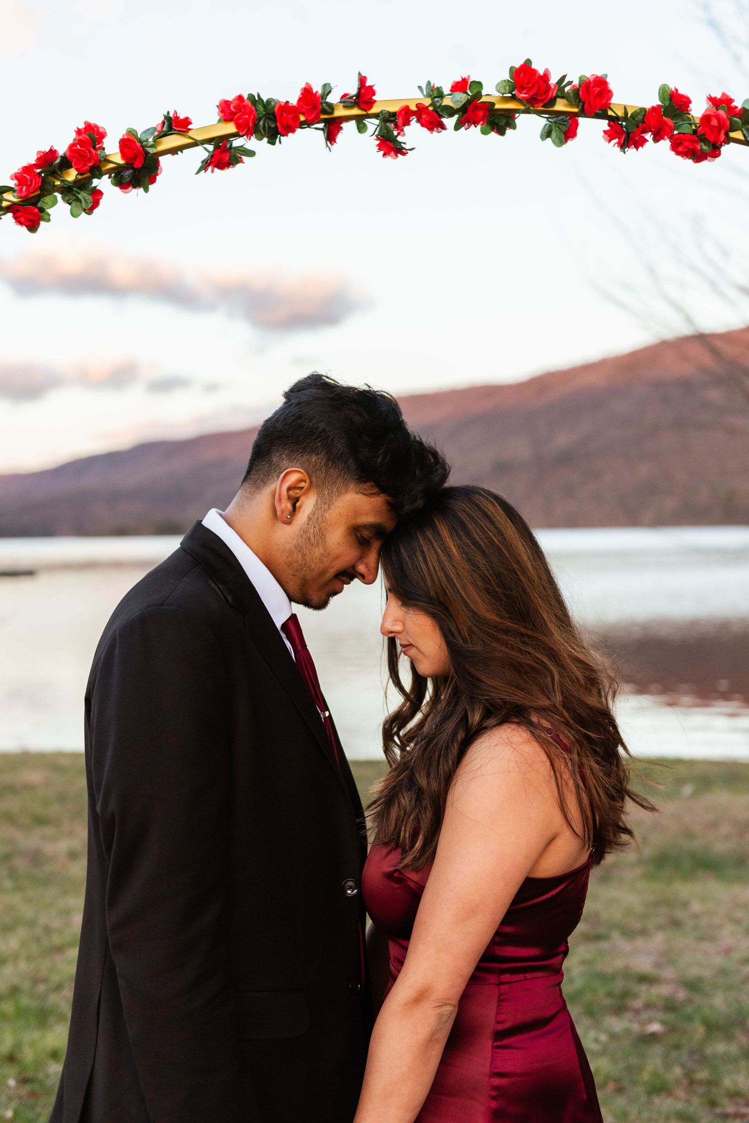 A romantic couple shares an intimate moment by a lake at sunset under an arch decorated with red flowers.