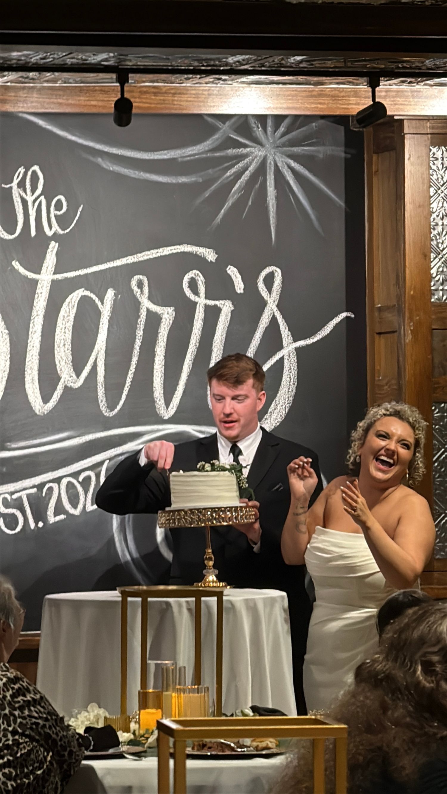A joyful wedding reception moment with a couple at their cake-cutting ceremony in front of a decorative chalkboard wall.