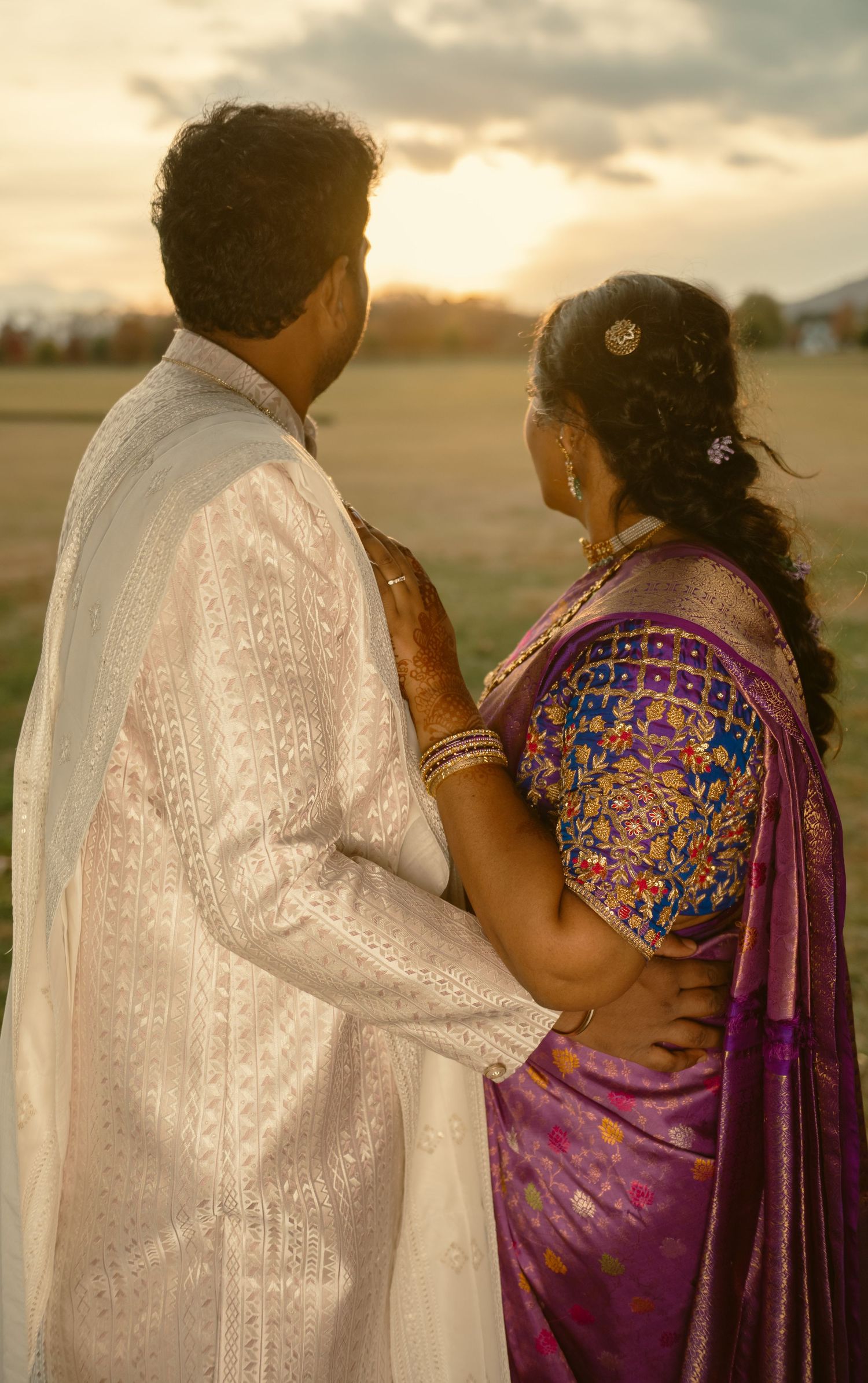 A couple in traditional Indian attire embraces on a beach at sunset, with the woman wearing a vibrant purple sari.