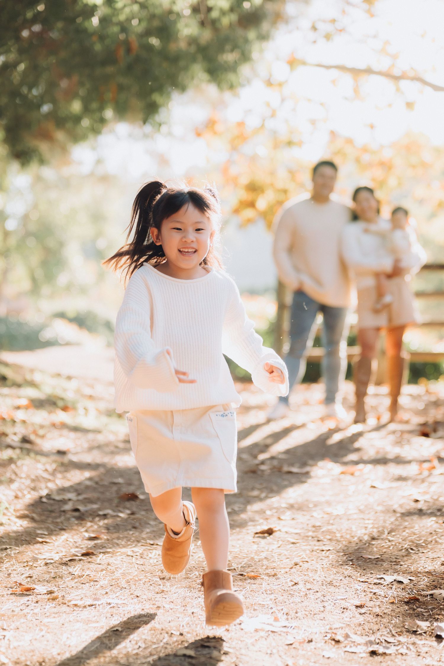A young child in a white dress runs playfully through a sun-dappled outdoor path during autumn.