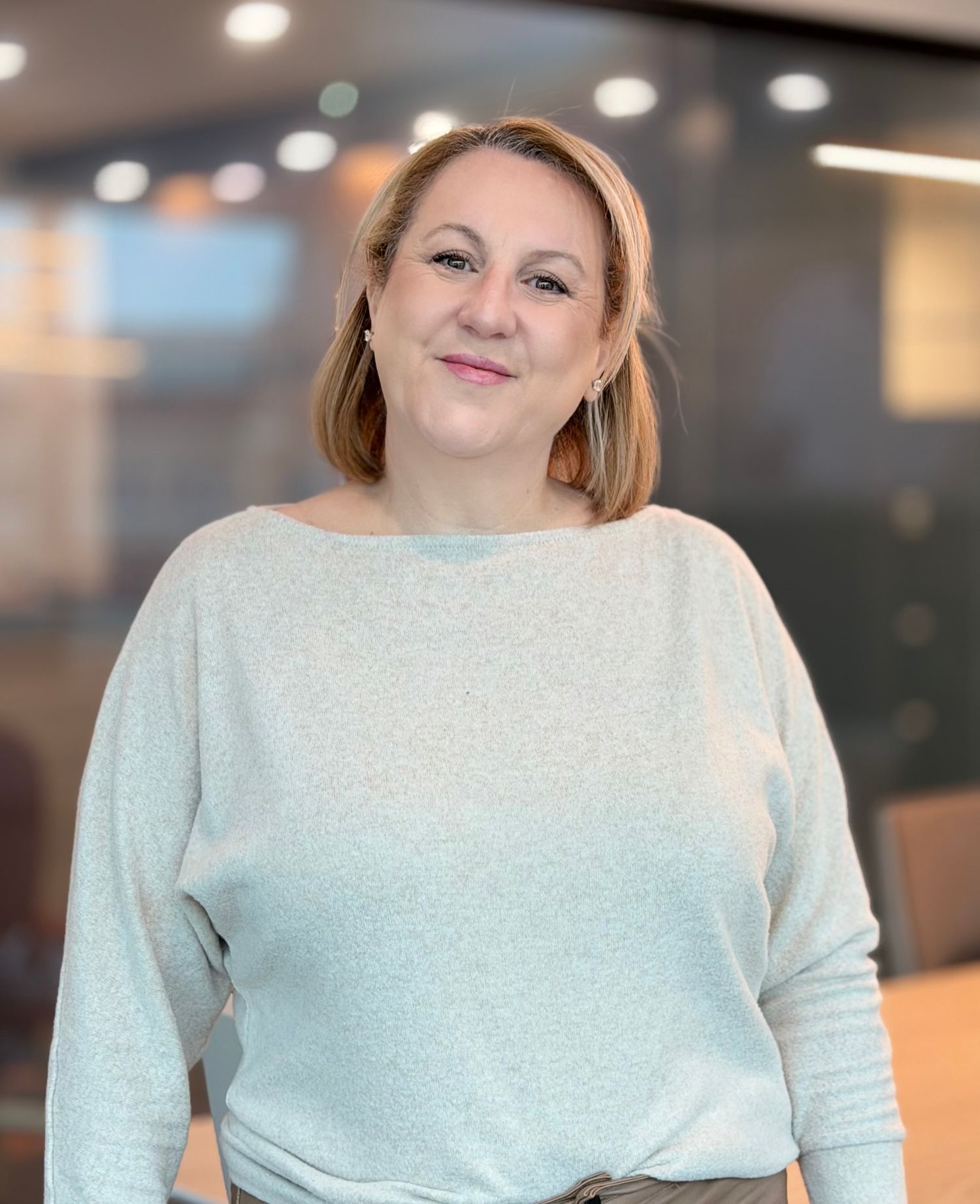 Smiling woman with blonde hair wearing a light blue sweater standing in a modern office environment.