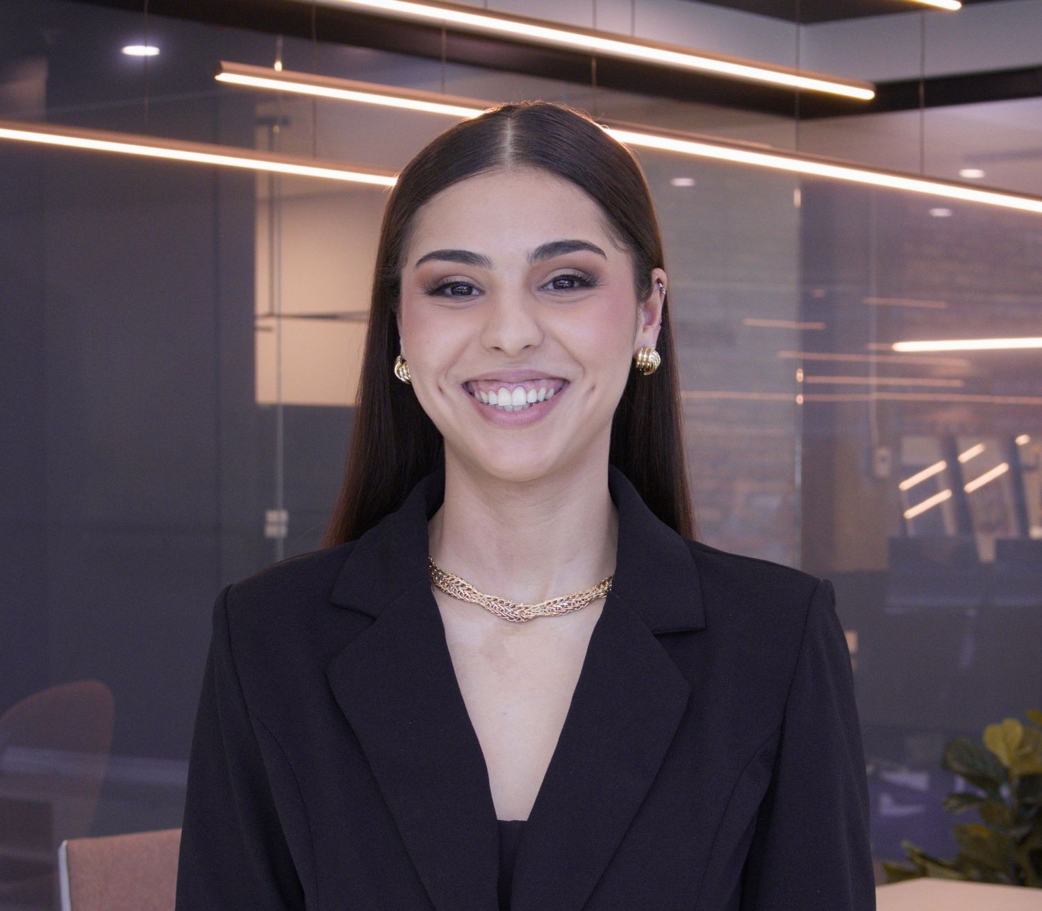 Professional young woman in black blazer smiling confidently in a modern office environment.