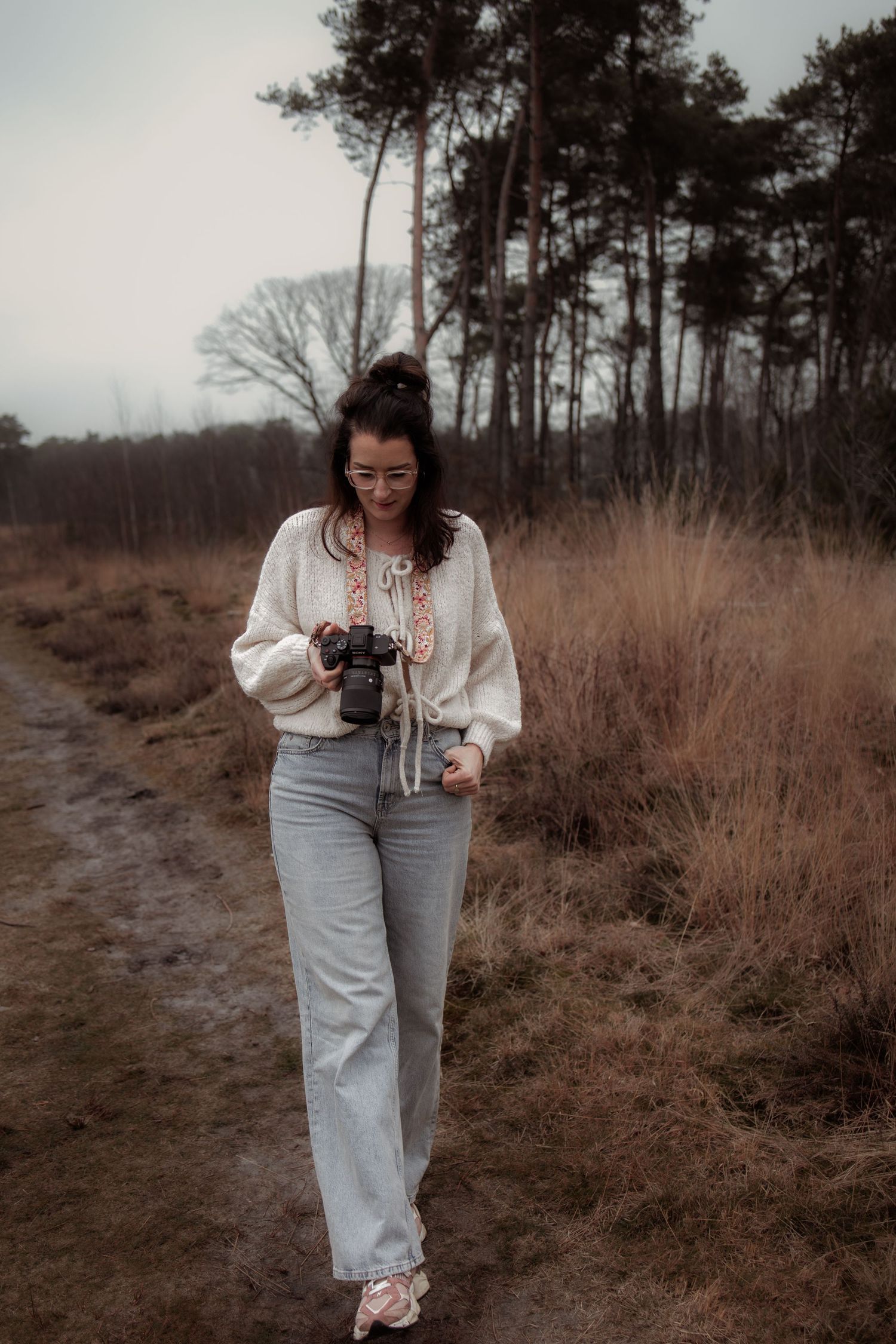 A person in a white top and light pants walks along a nature trail surrounded by brown grass and pine trees.