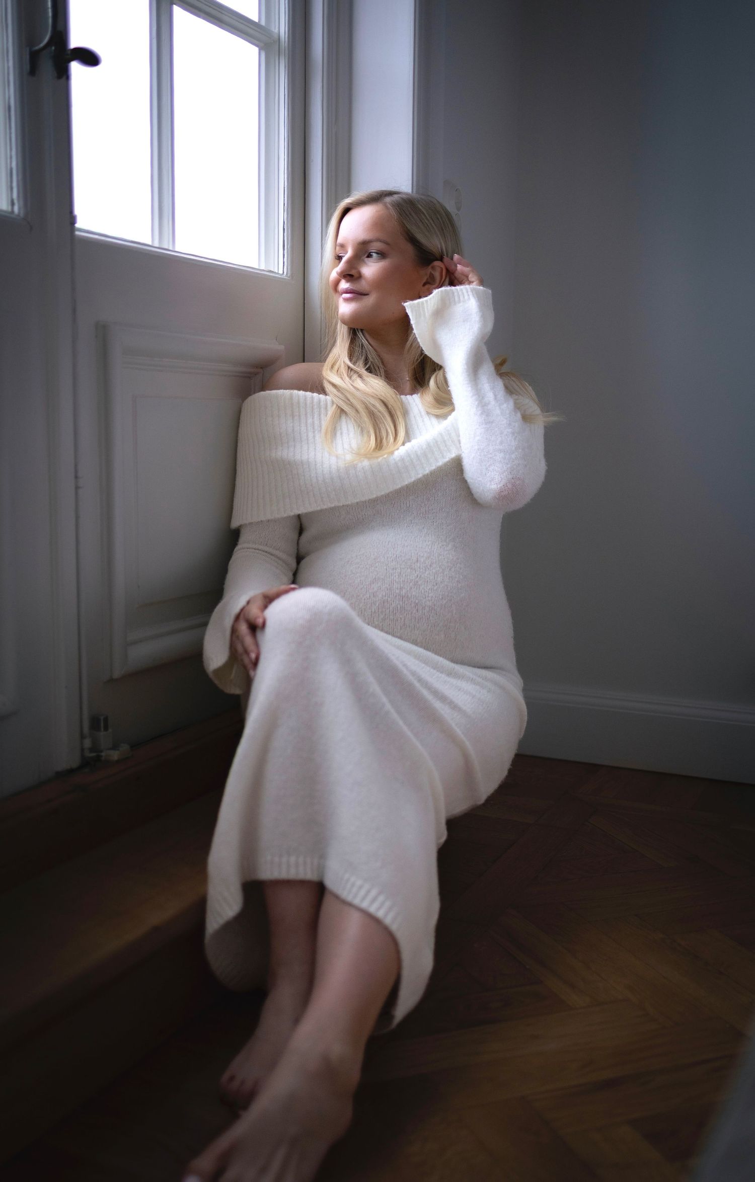 gravid kvinna sitter på ett trappsteg lutande mot en dörr med fönster som hon kollar ut genom. på sig har hon en vit stickad klänning